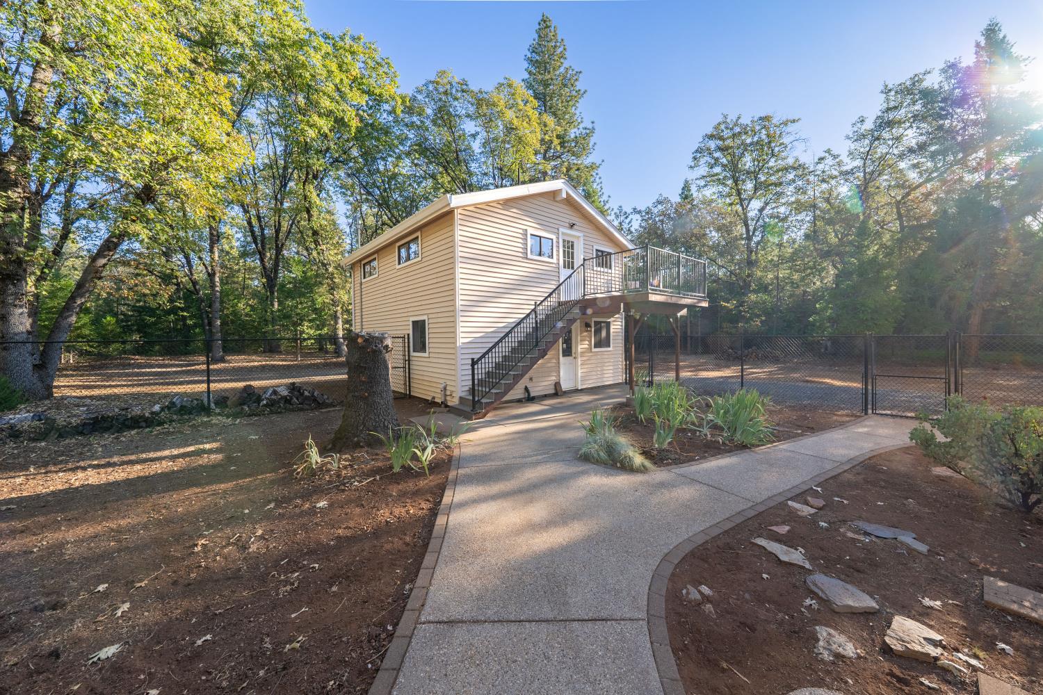 Beige raised home with an elevated deck and outdoor stairs, set among mature trees beside a curved walkway.
