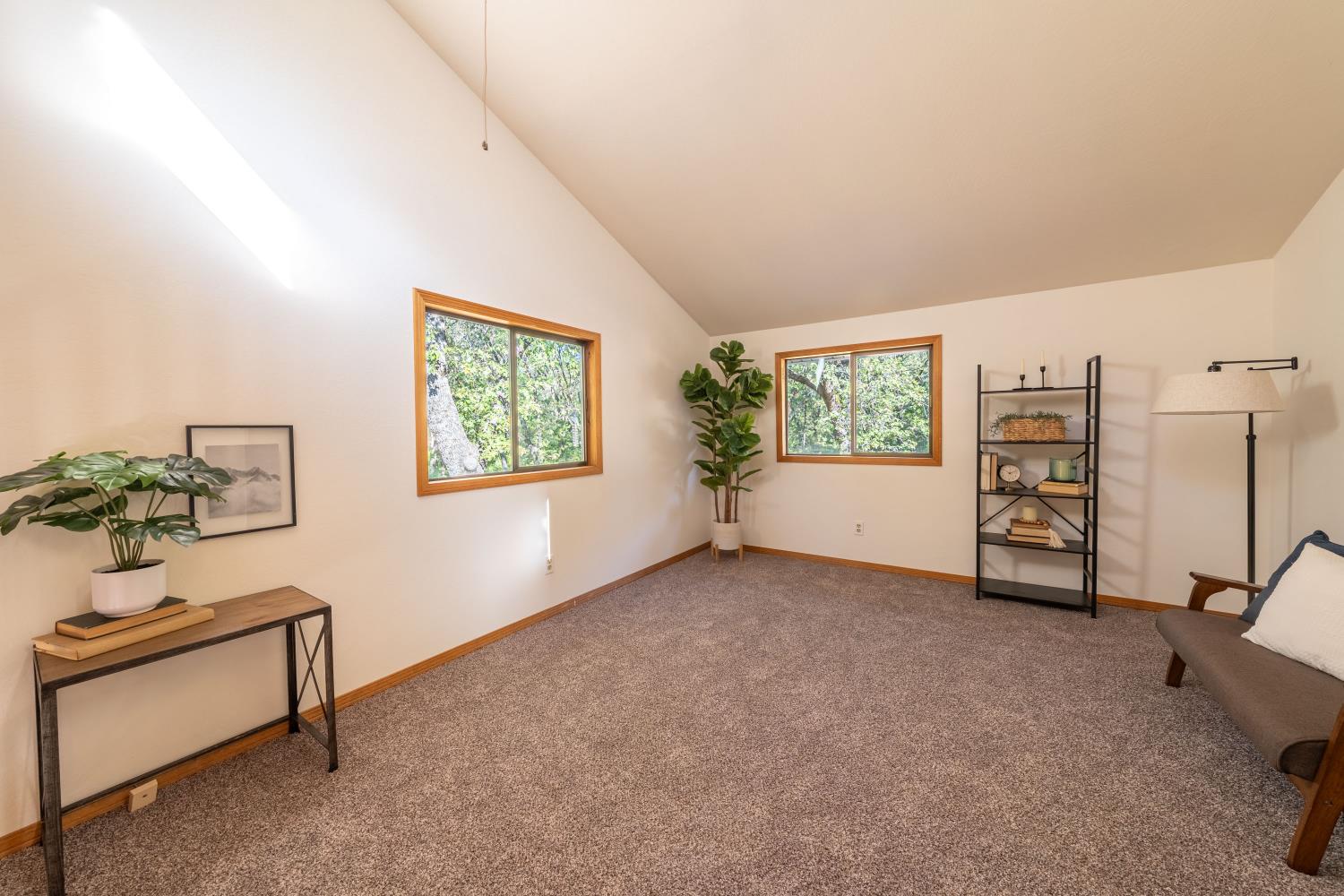 Carpeted room with a vaulted ceiling, two tree-facing windows, and simple staged furnishings.