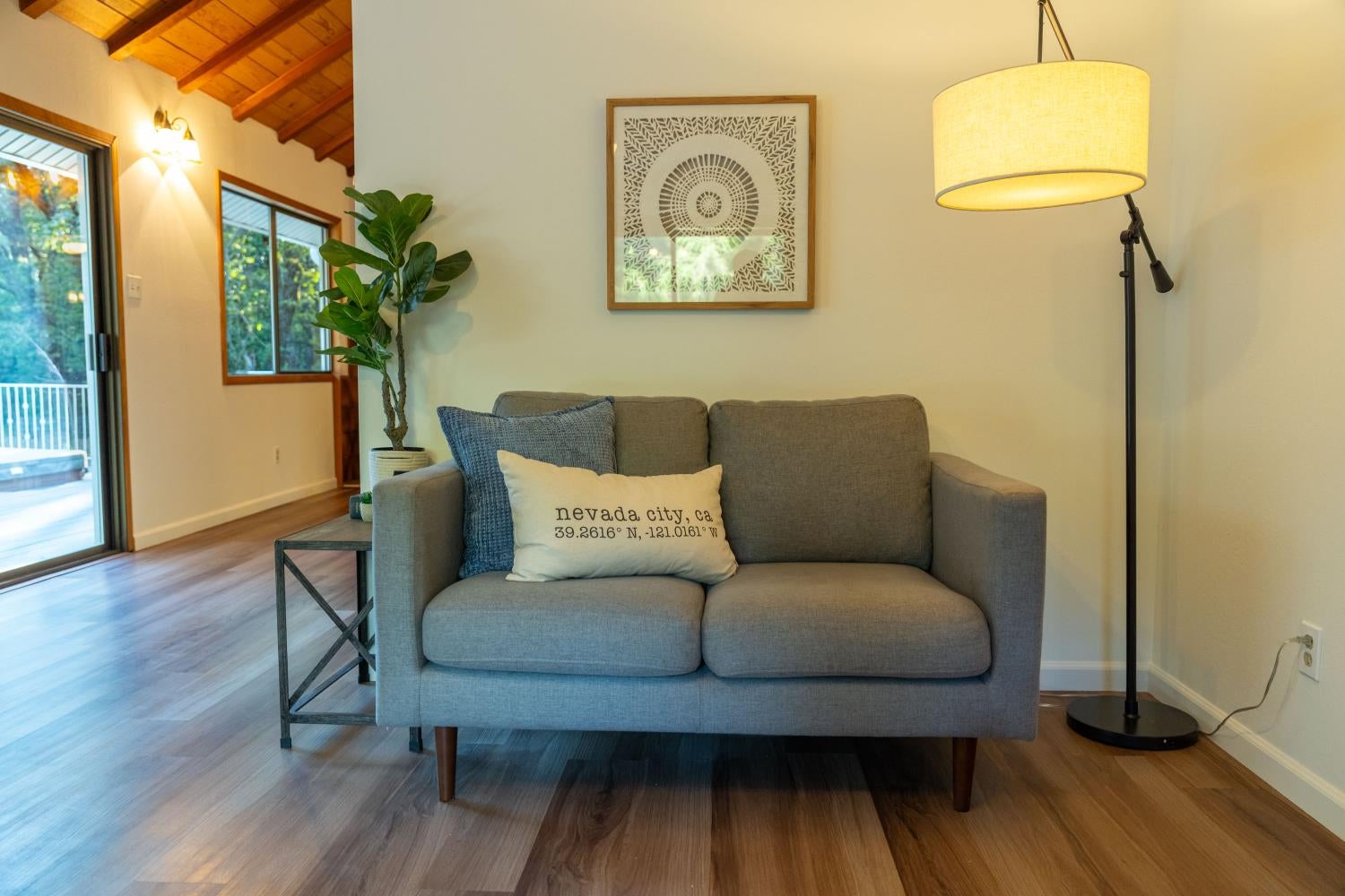 Staged living room with gray loveseat, Nevada City pillow, tall floor lamp, and vaulted wood ceiling.