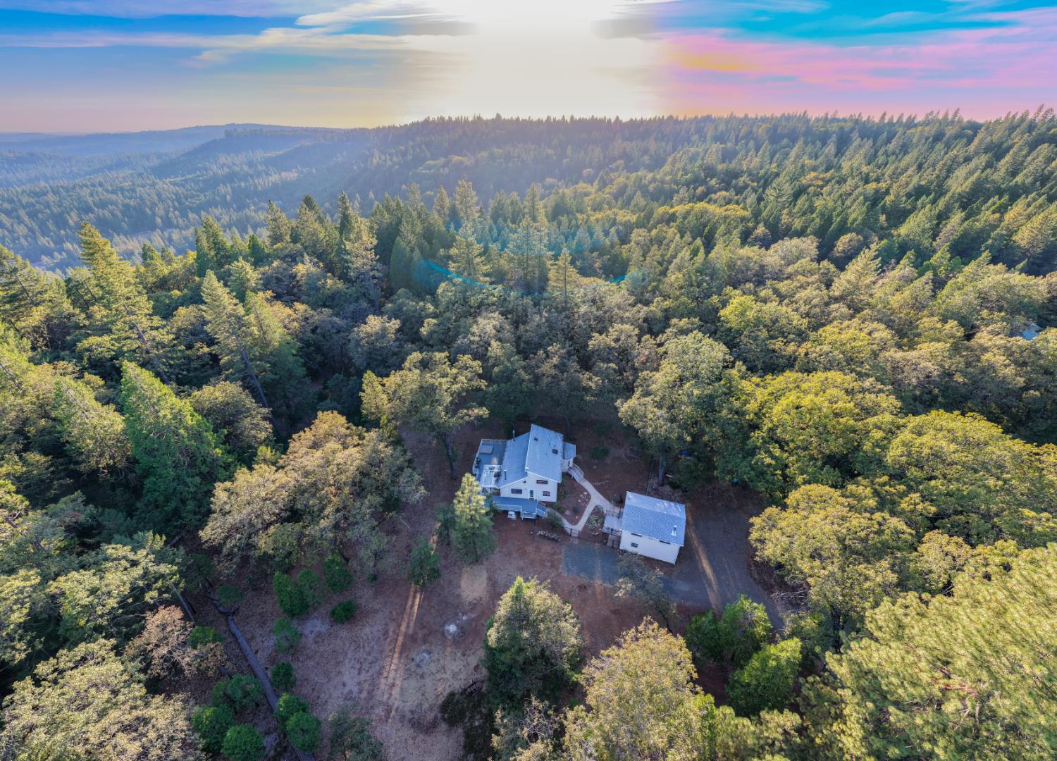 Aerial view of a home and outbuilding amid dense forest, with wooded hills on the horizon.