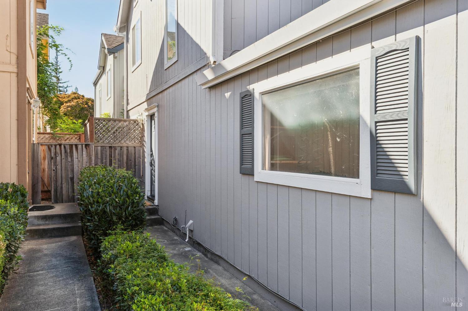 Gray home’s side yard with a shuttered window, concrete walkway, shrubs, and wooden fence.