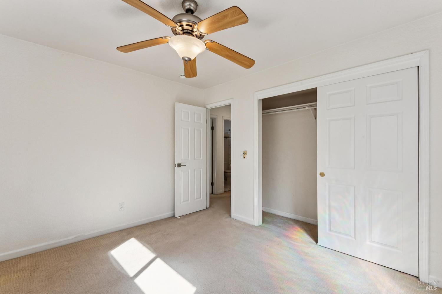 Empty bedroom with beige carpet, a wood-blade ceiling fan, and an open closet.
