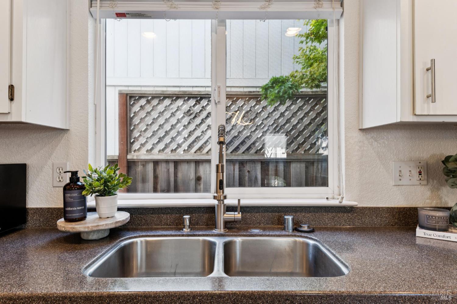 Kitchen sink with a tall pull-down faucet, dark countertop, white cabinets, and a window overlooking a fenced yard.