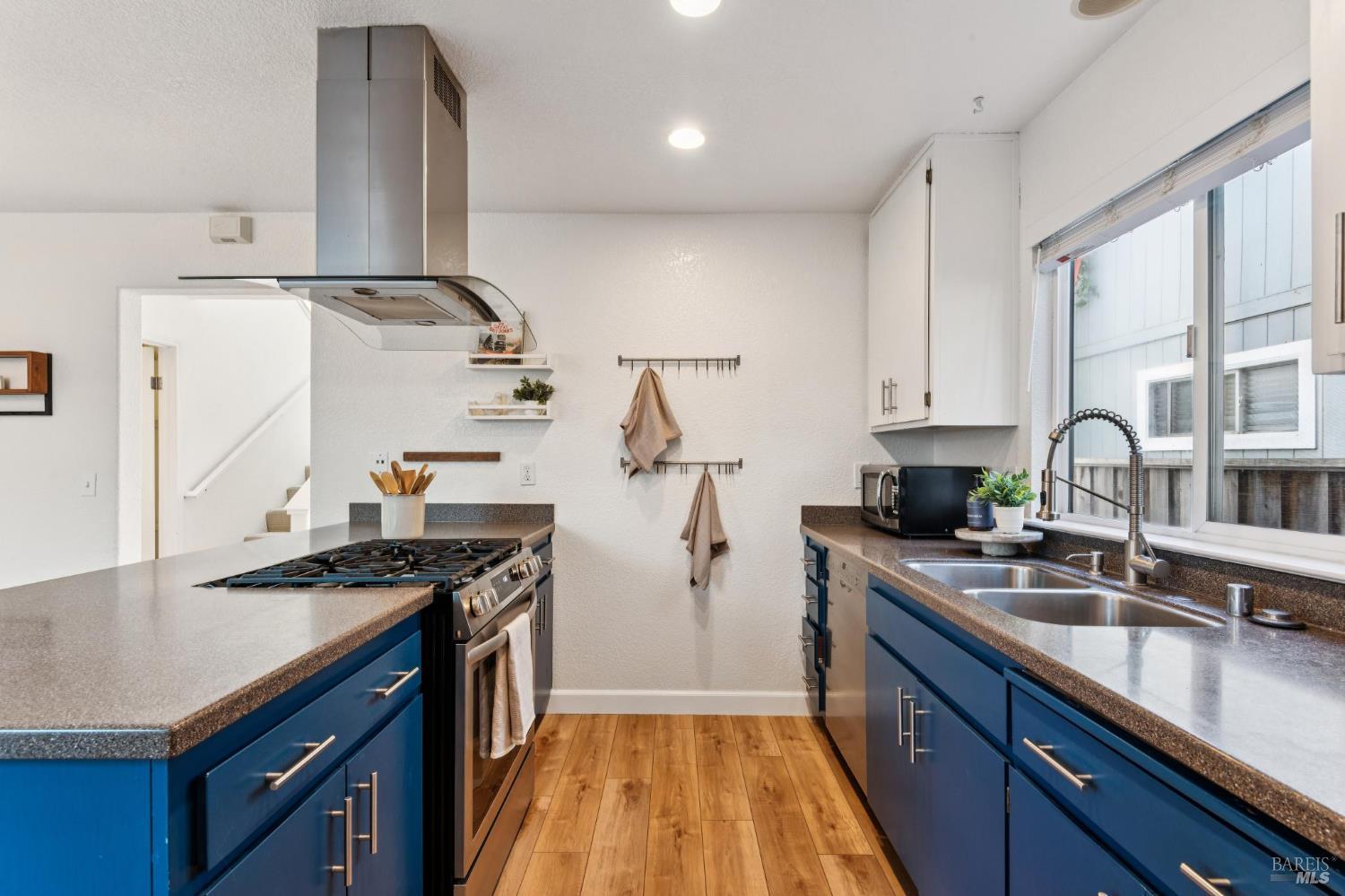 Galley kitchen with blue cabinets, wood floors, and a double-basin sink beneath a large window.