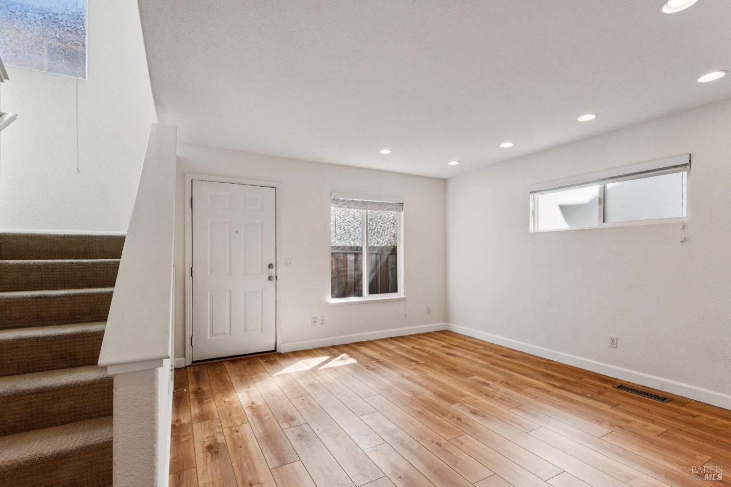 Vacant living room with warm wood floors, front door, windows, and carpeted staircase.