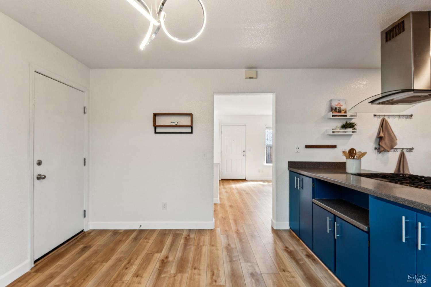 Kitchen with blue cabinets, dark countertops, and wood-look floors leading to an adjoining room.