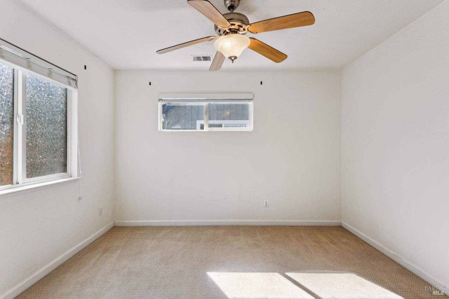 Empty bedroom with beige carpet, two windows, and a wood-bladed ceiling fan.