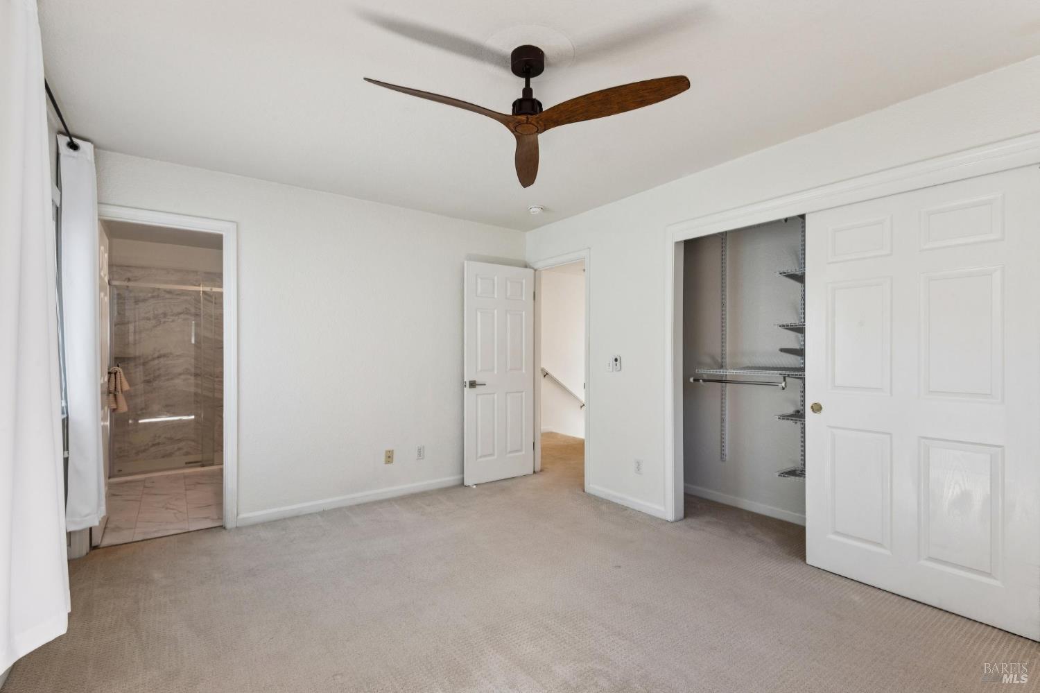 Empty carpeted bedroom with a wood ceiling fan, open wire-shelved closet, and adjoining tiled bathroom.