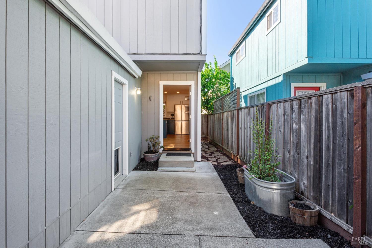 Side patio and concrete walkway leading to an open doorway, bordered by planters and a wooden fence.