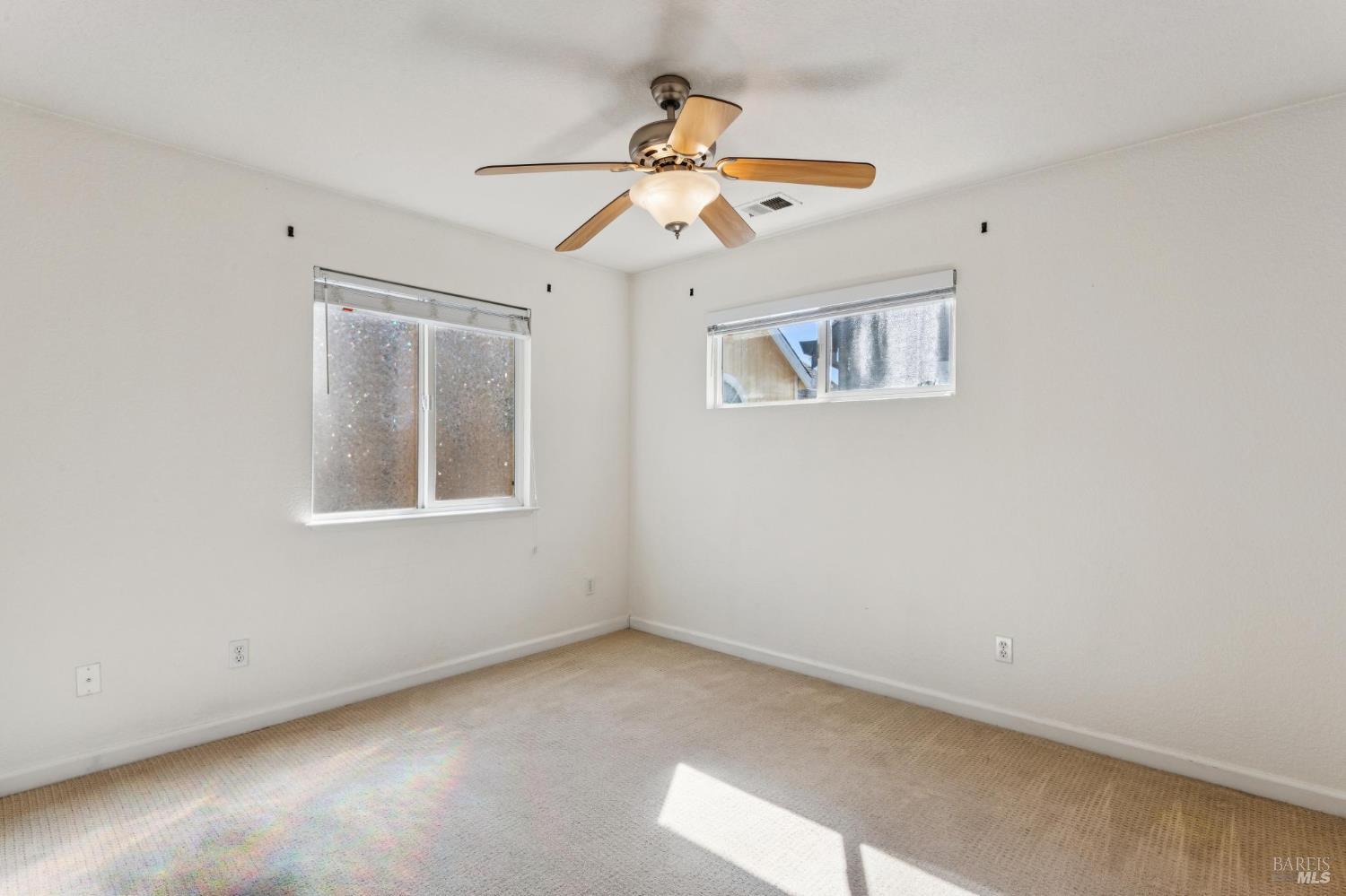 Empty bedroom with beige carpet, two windows, and a ceiling fan with a light.