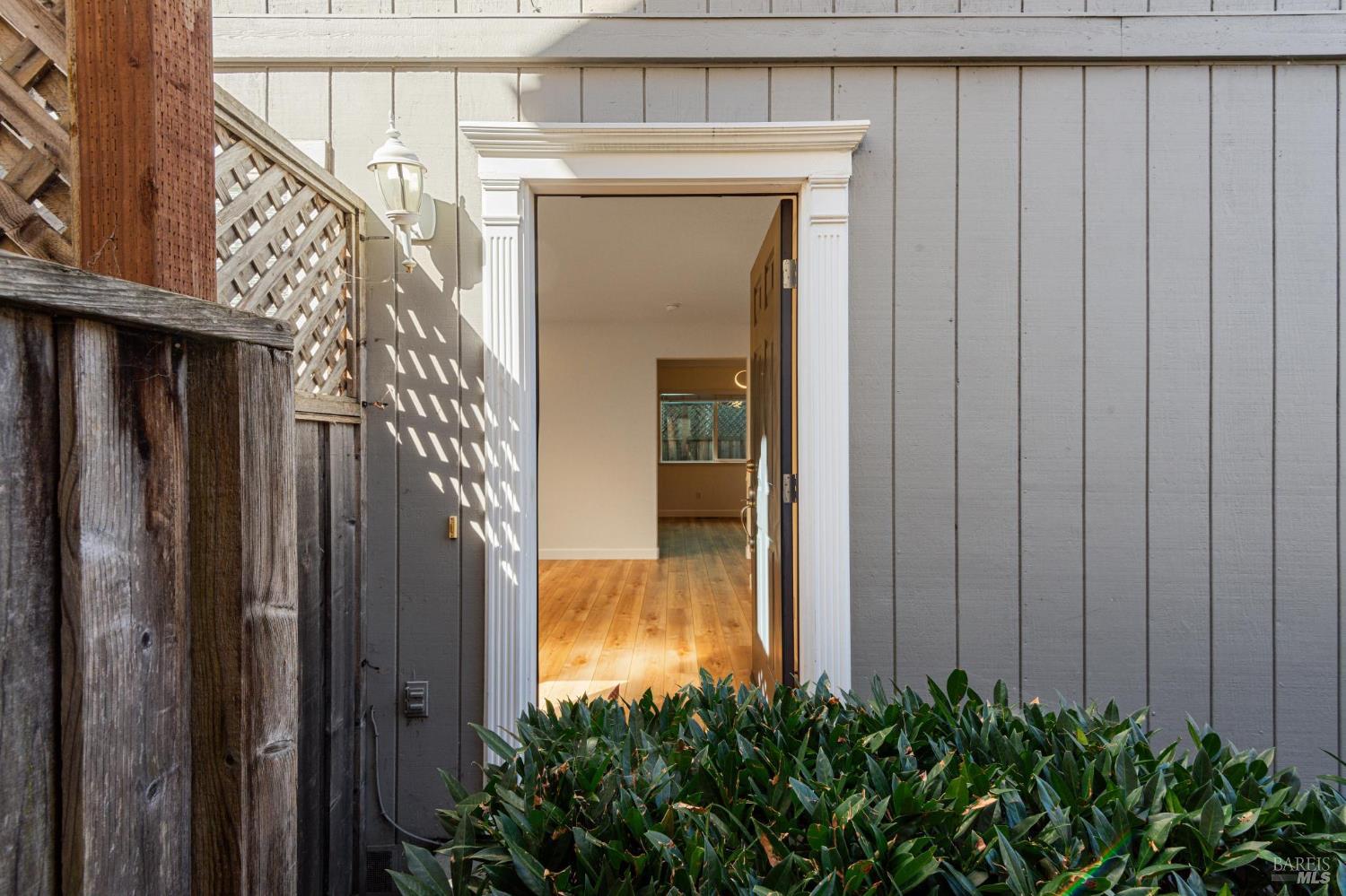 Open, white-trimmed entry in gray siding, with a weathered fence and leafy shrubs.