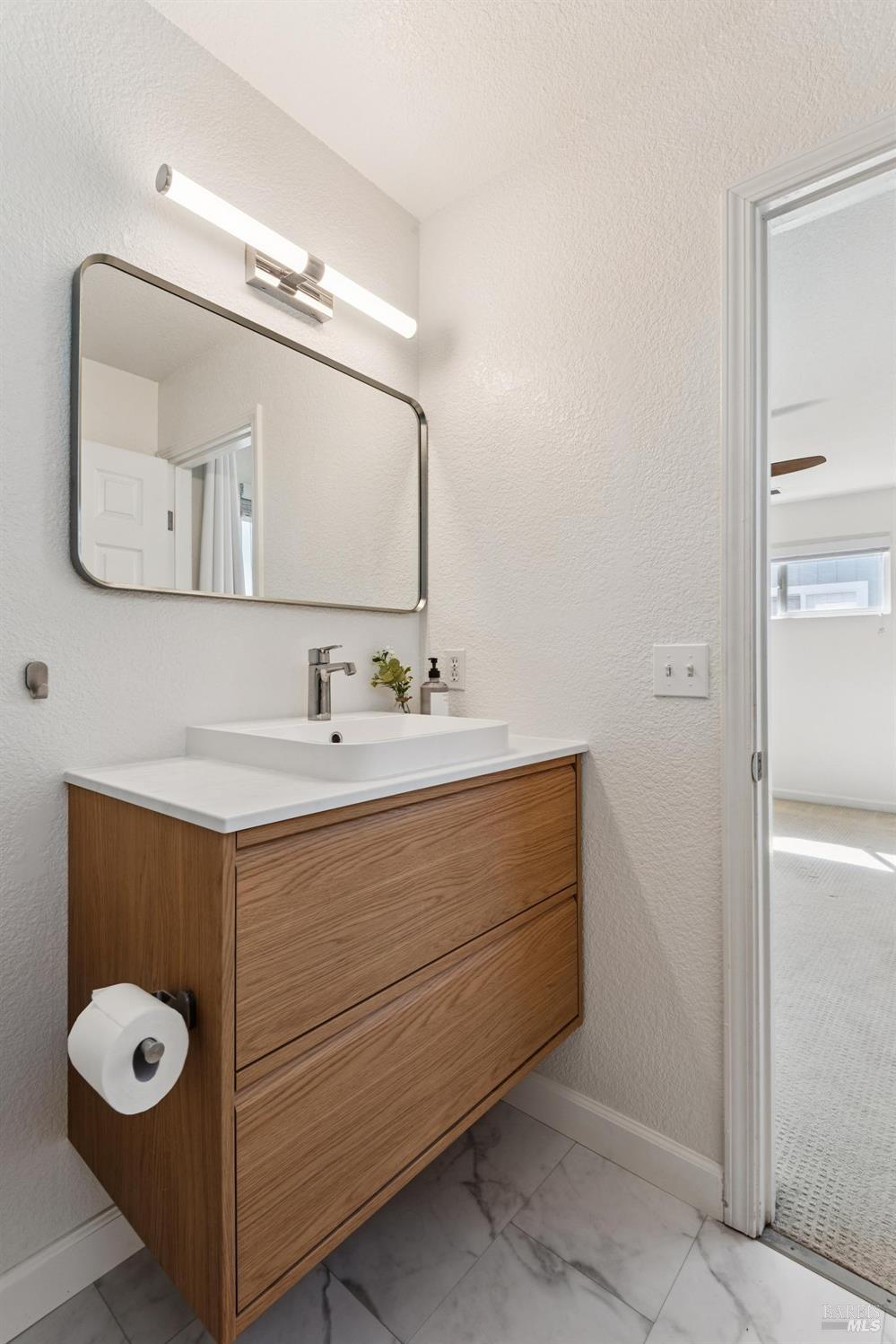 Bathroom vanity with a wood-grain cabinet, white vessel sink, large mirror, and marble-look floor.