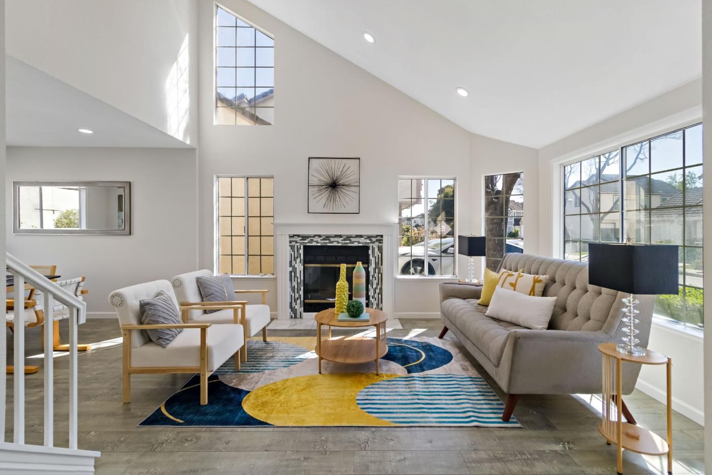Staged living room with a vaulted ceiling, tall windows, tiled fireplace, and colorful geometric rug.