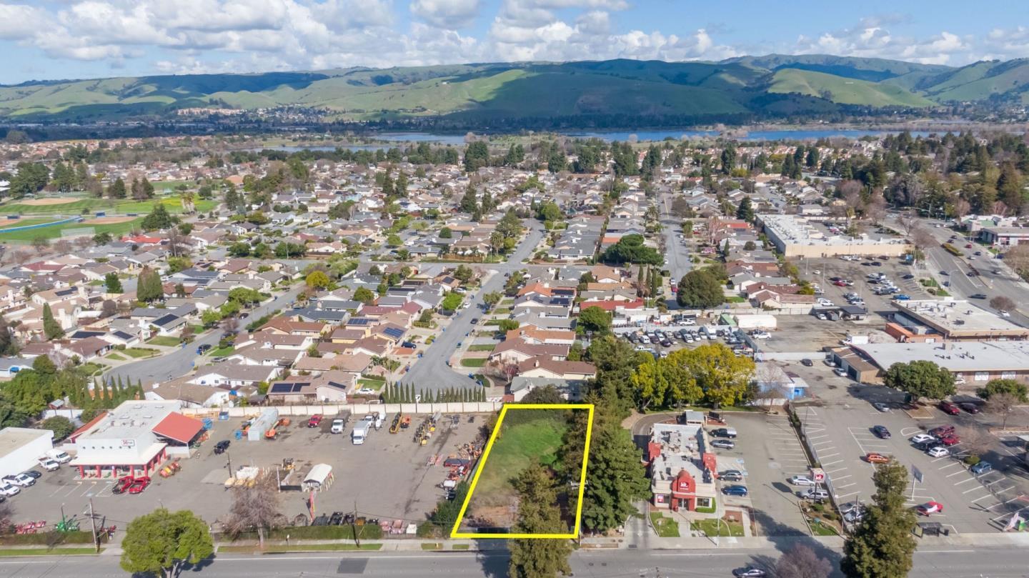Vacant parcel outlined in yellow, surrounded by homes and businesses with hills and water beyond.