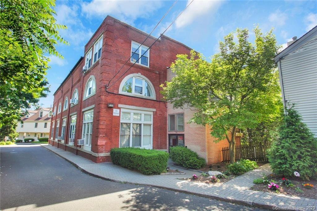 Red brick corner building with arched windows, white trim, and shrubs along the sidewalk.
