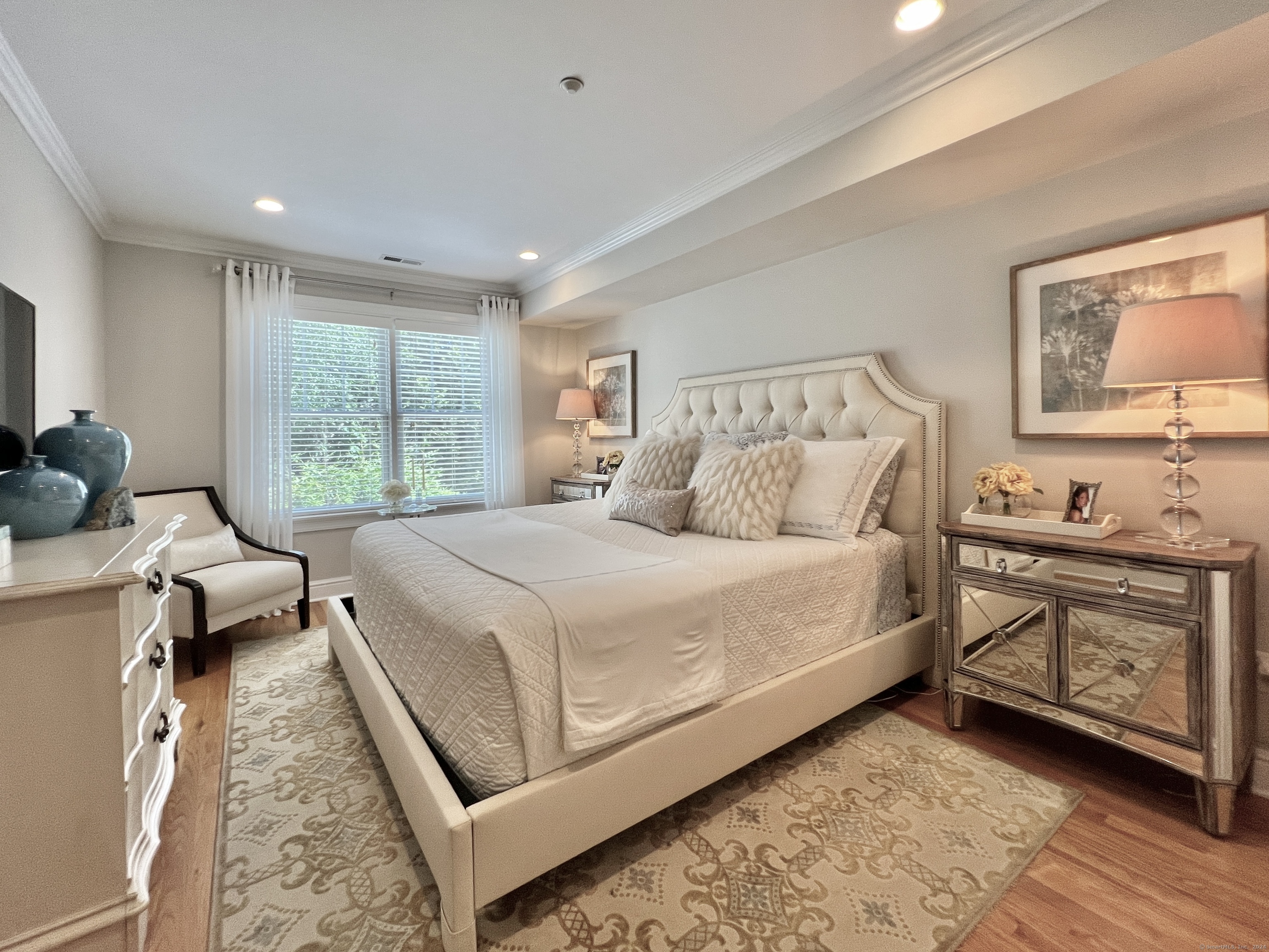 Furnished bedroom with a cream tufted bed, patterned rug, and large window with white blinds.