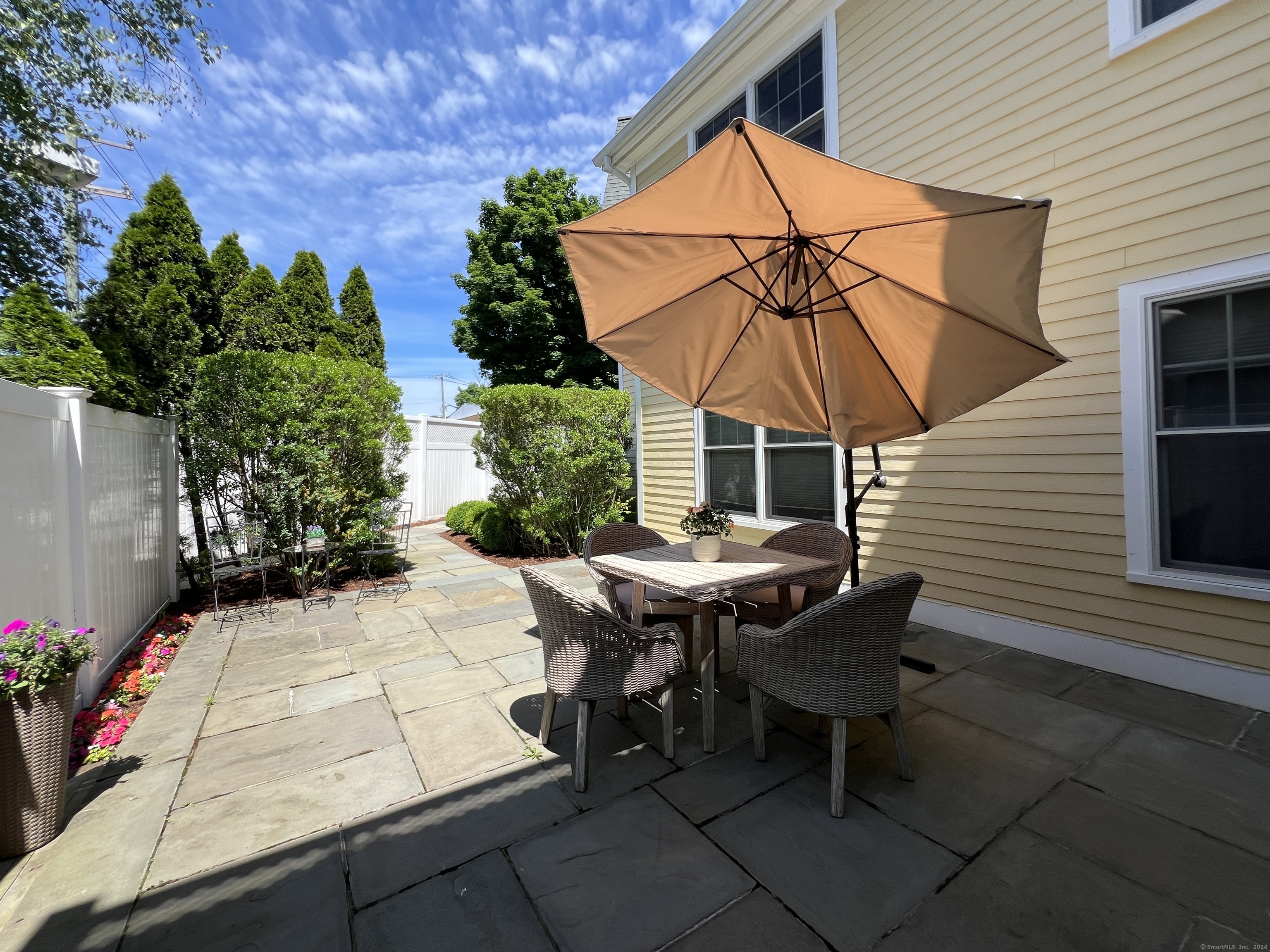 Backyard patio with wicker dining set beneath a tan umbrella, bordered by stone pavers and leafy shrubs.