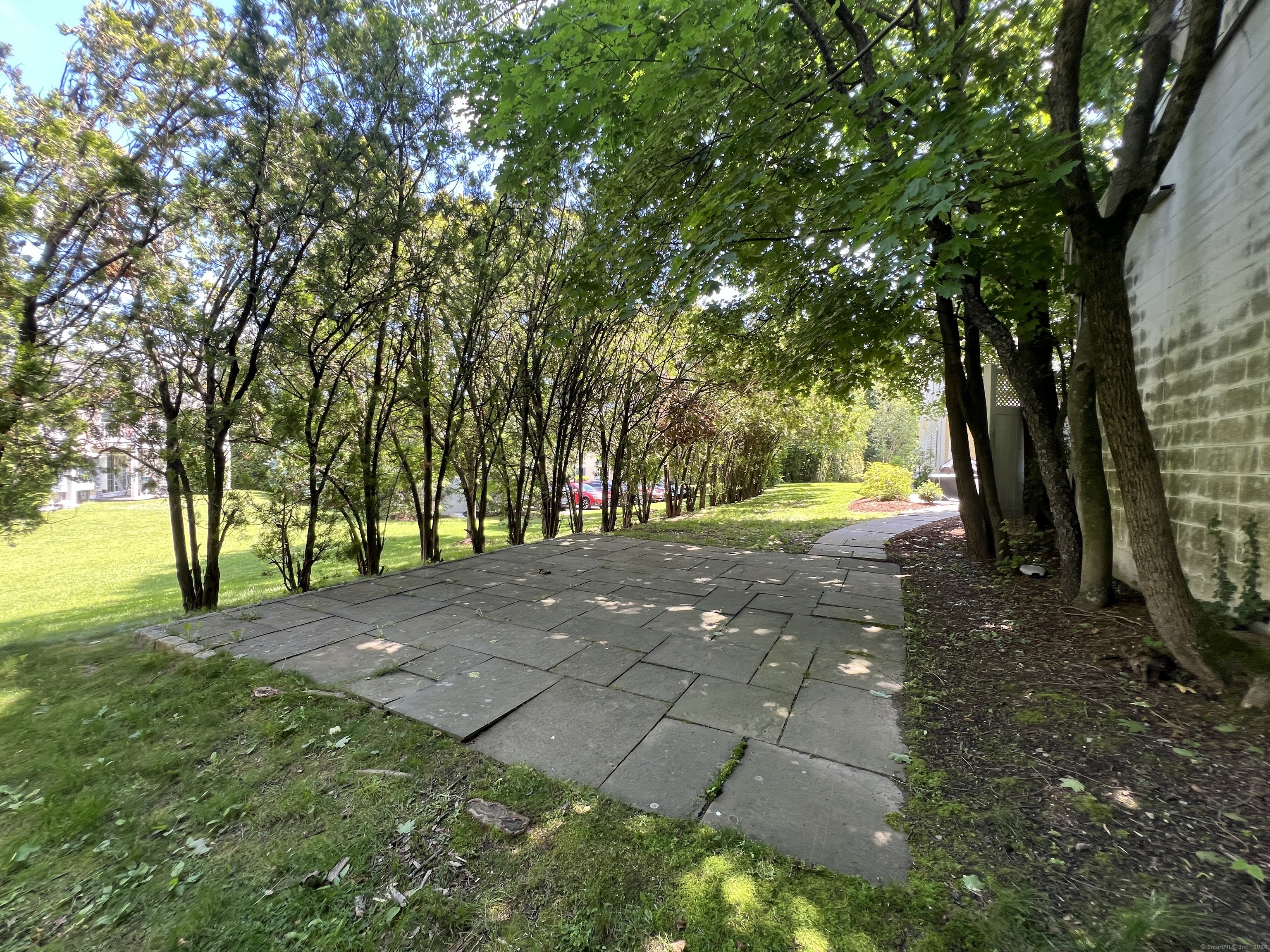 Shaded stone patio beside a building, surrounded by mature trees and lawn.