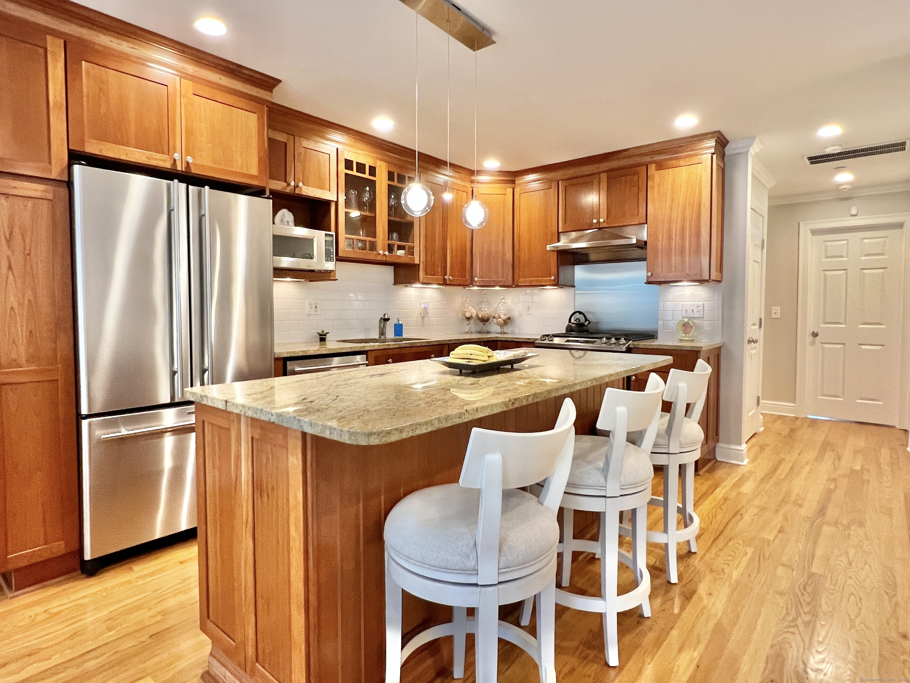 Kitchen with a granite island, four white stools, wood cabinets, and stainless steel appliances.