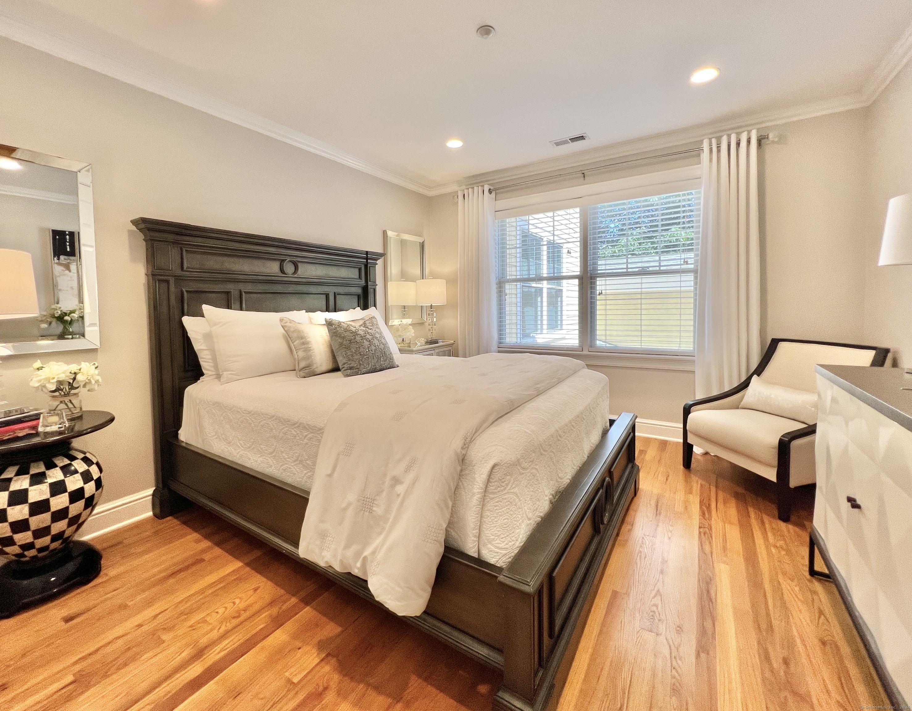 Staged bedroom with a dark wood bed, white bedding, hardwood floors, and a chair by the window.