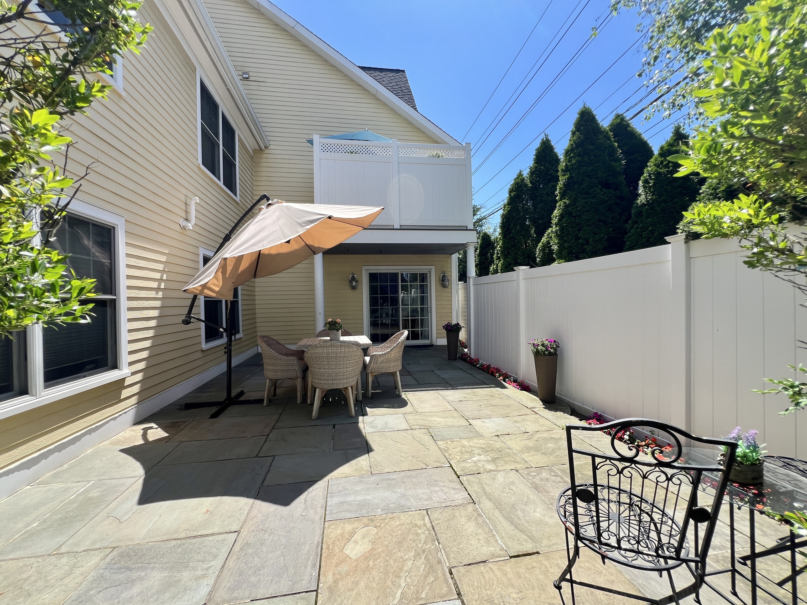 Stone patio with umbrella-shaded dining set, white privacy fence, and an upper deck.