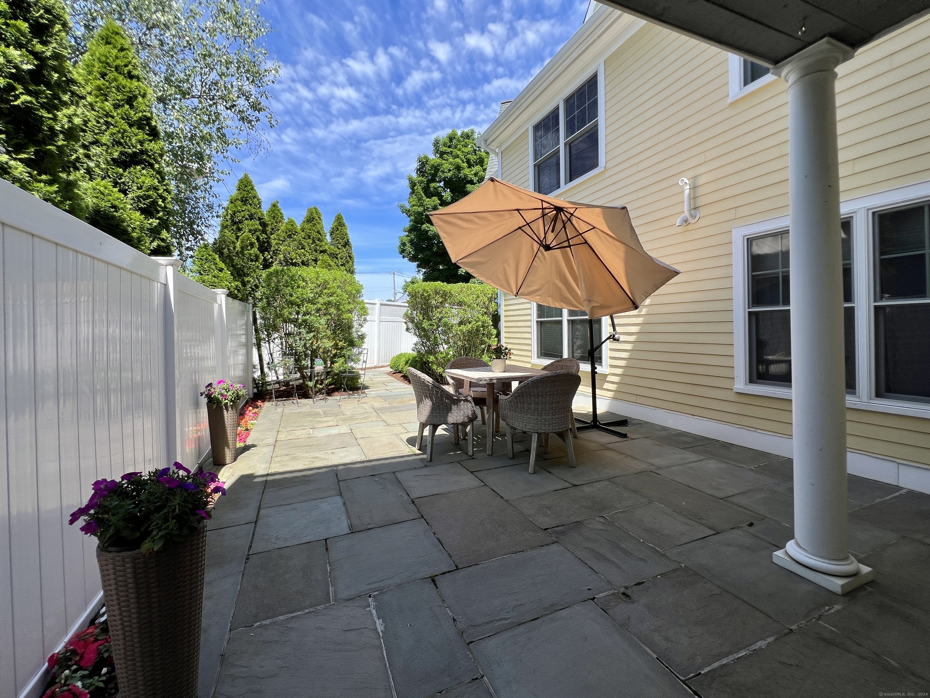 Stone patio with wicker dining set and tan umbrella beside a yellow house.