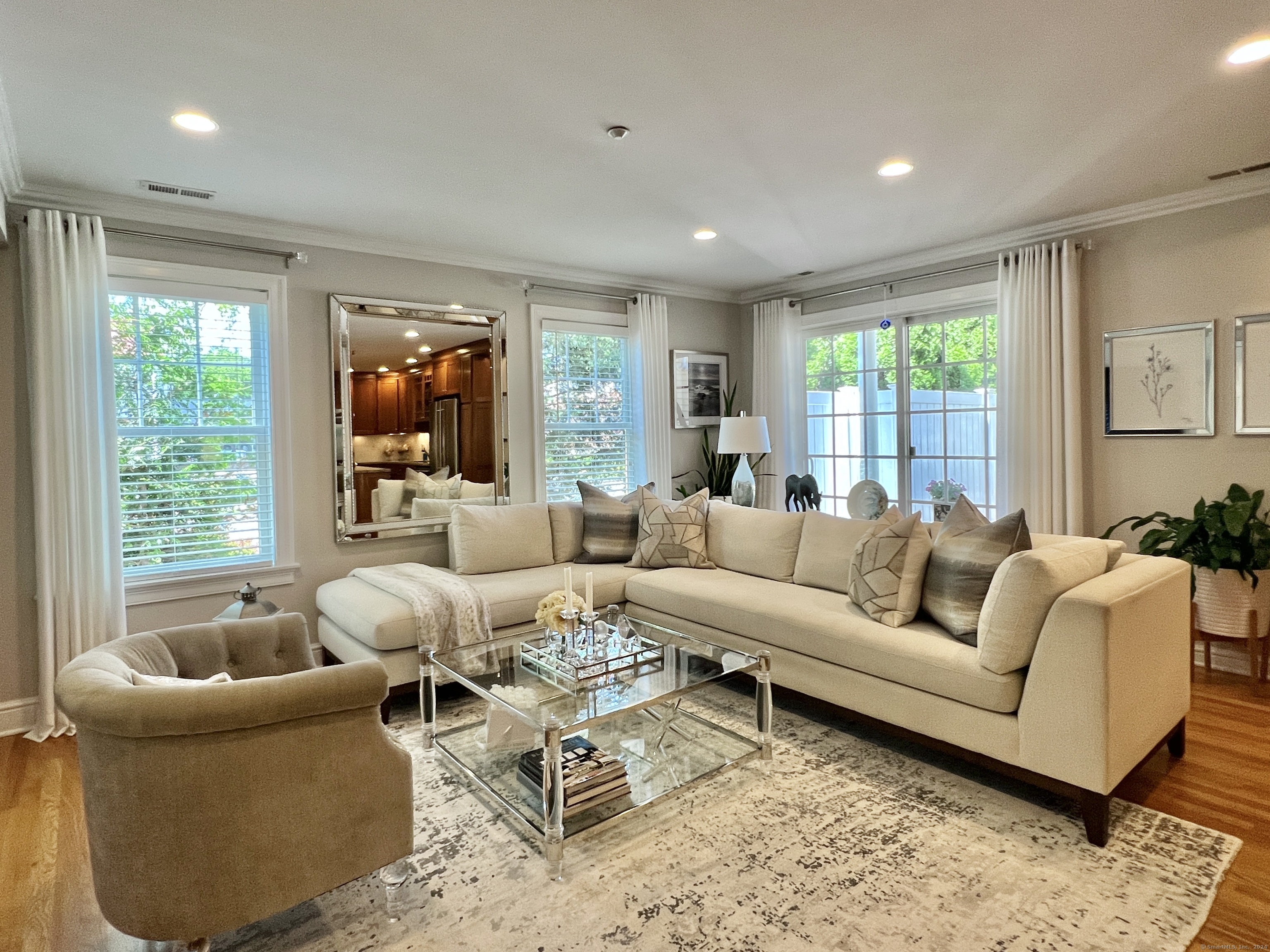Furnished living room with a cream sectional, glass coffee table, patterned rug, and windows overlooking greenery.