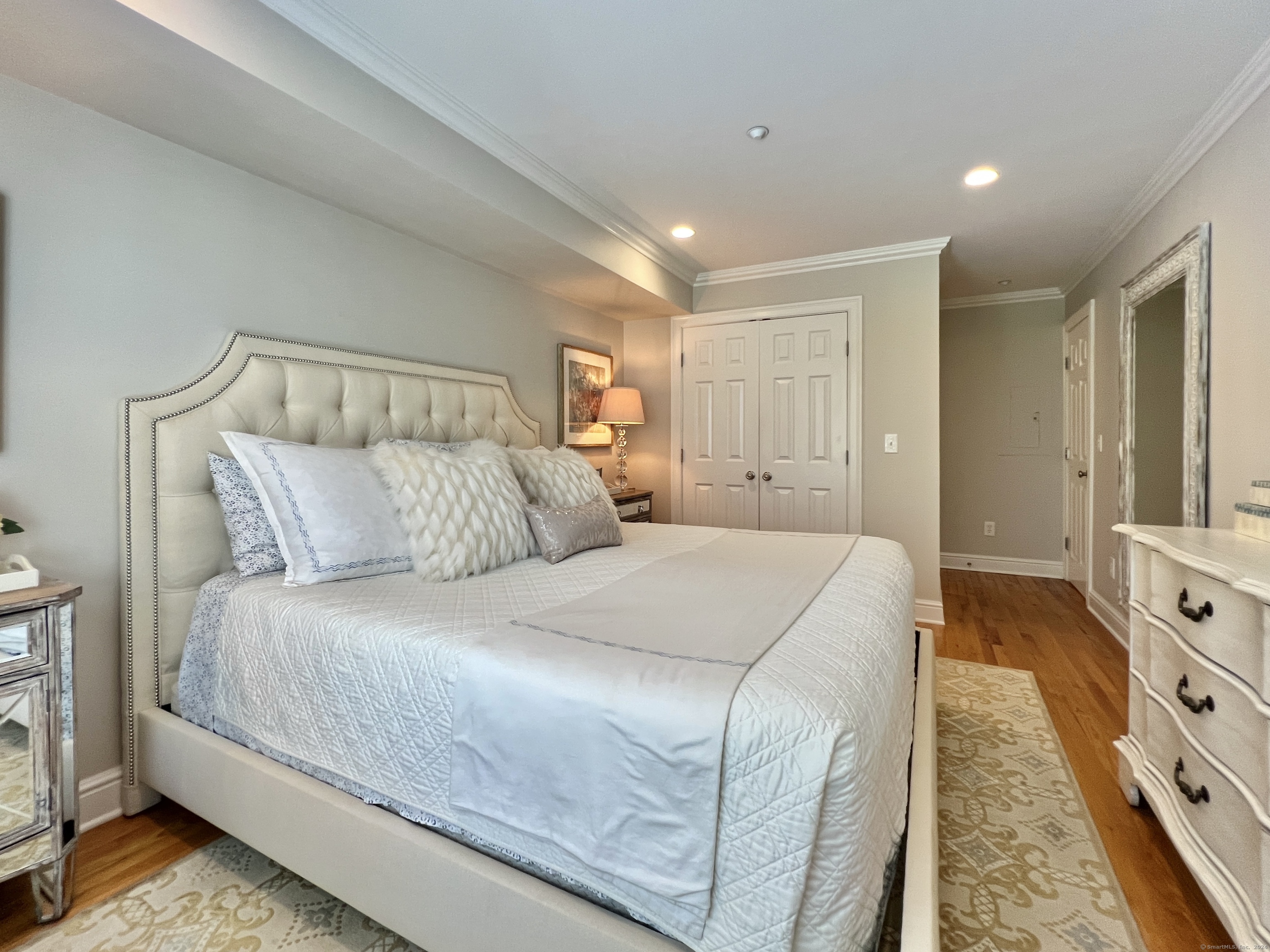 Bedroom with a large cream tufted bed, white quilted bedding, hardwood floors, and patterned rugs.