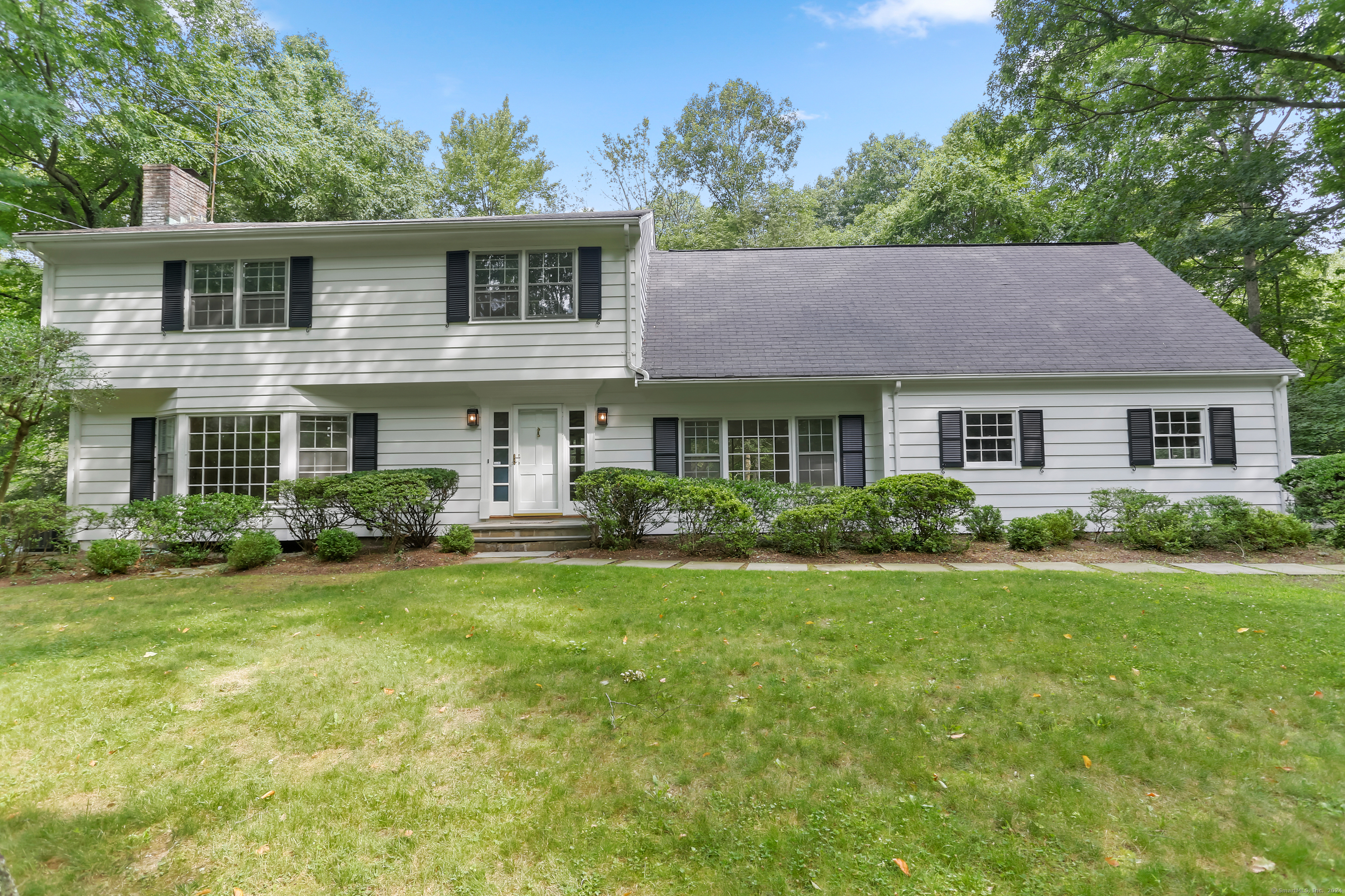 White two-story house with black shutters, a broad gray roof, and a lawn shaded by mature trees.