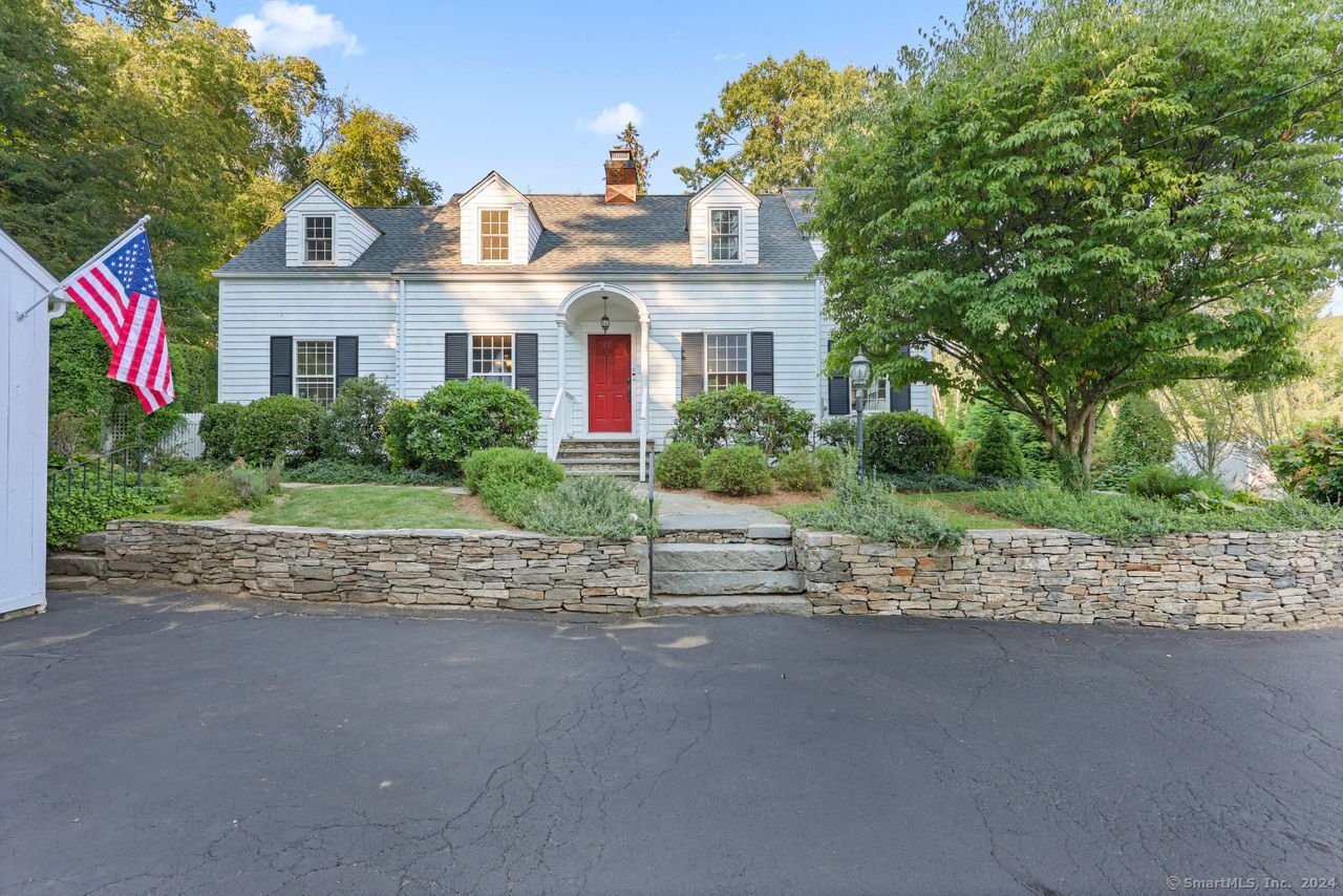 White house with three dormers and a red front door behind a stone retaining wall.