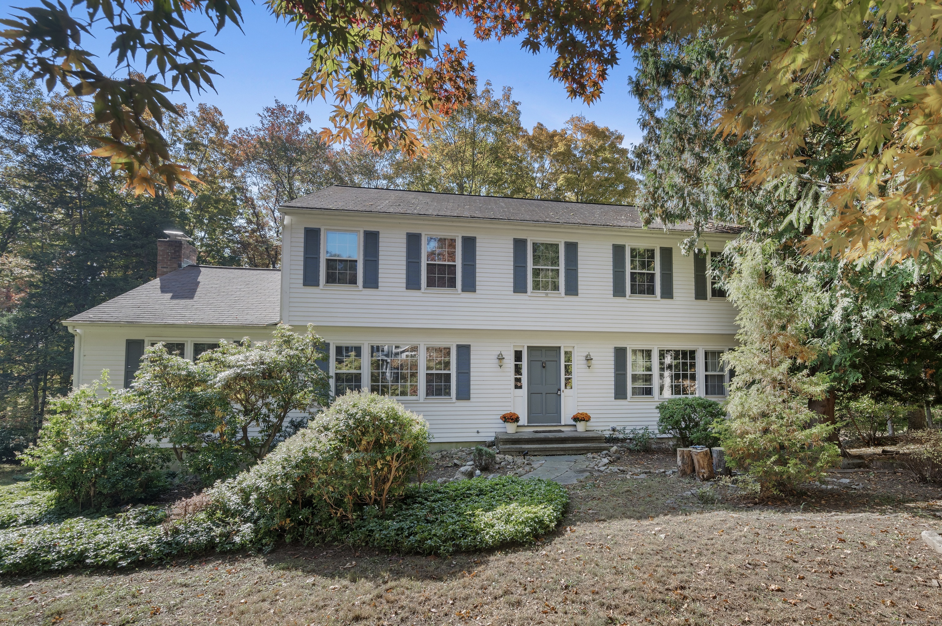 White two-story house with blue shutters, framed by mature trees in autumn.