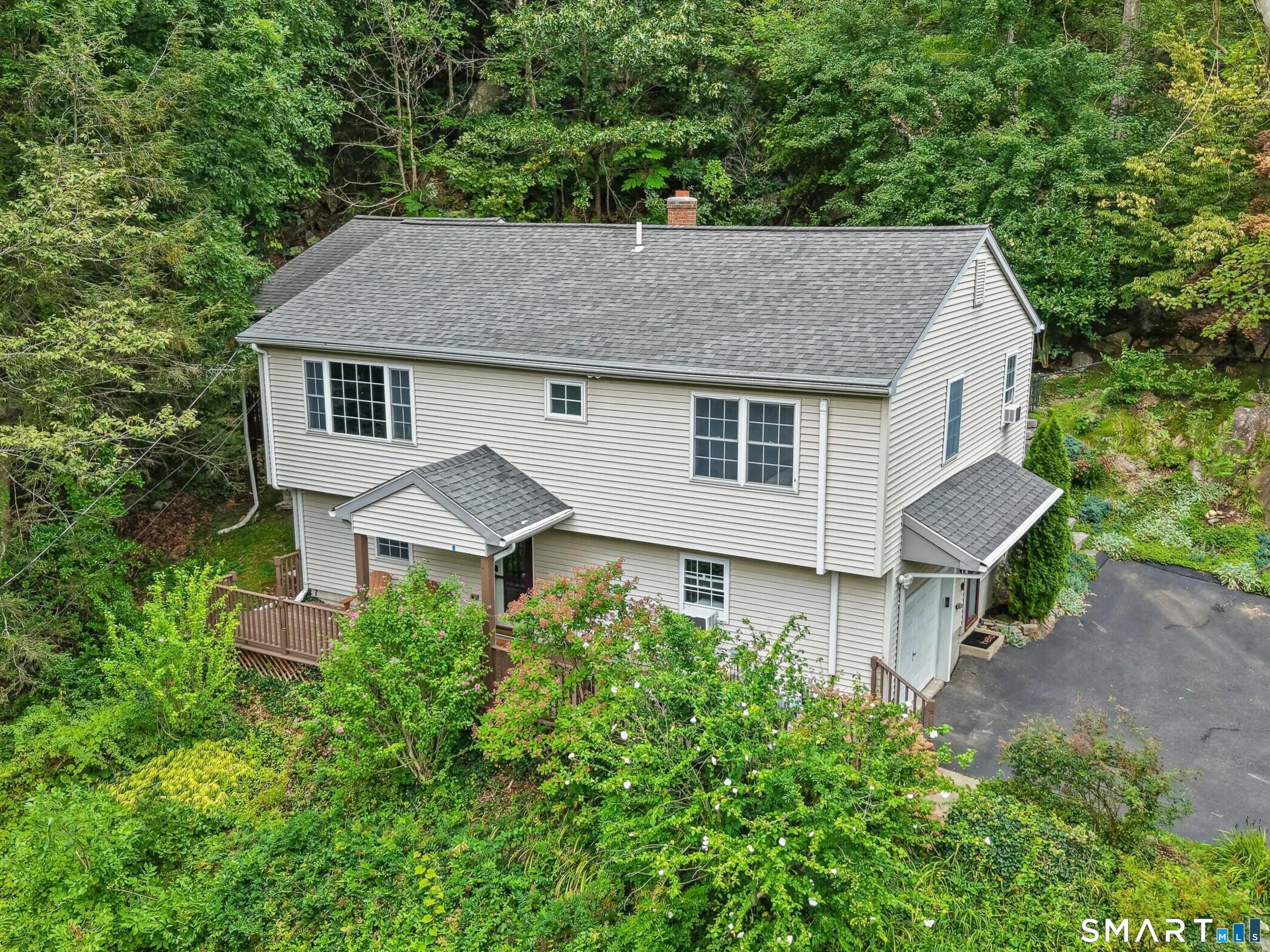 Two-story light-sided house surrounded by trees, with a small deck and paved driveway.