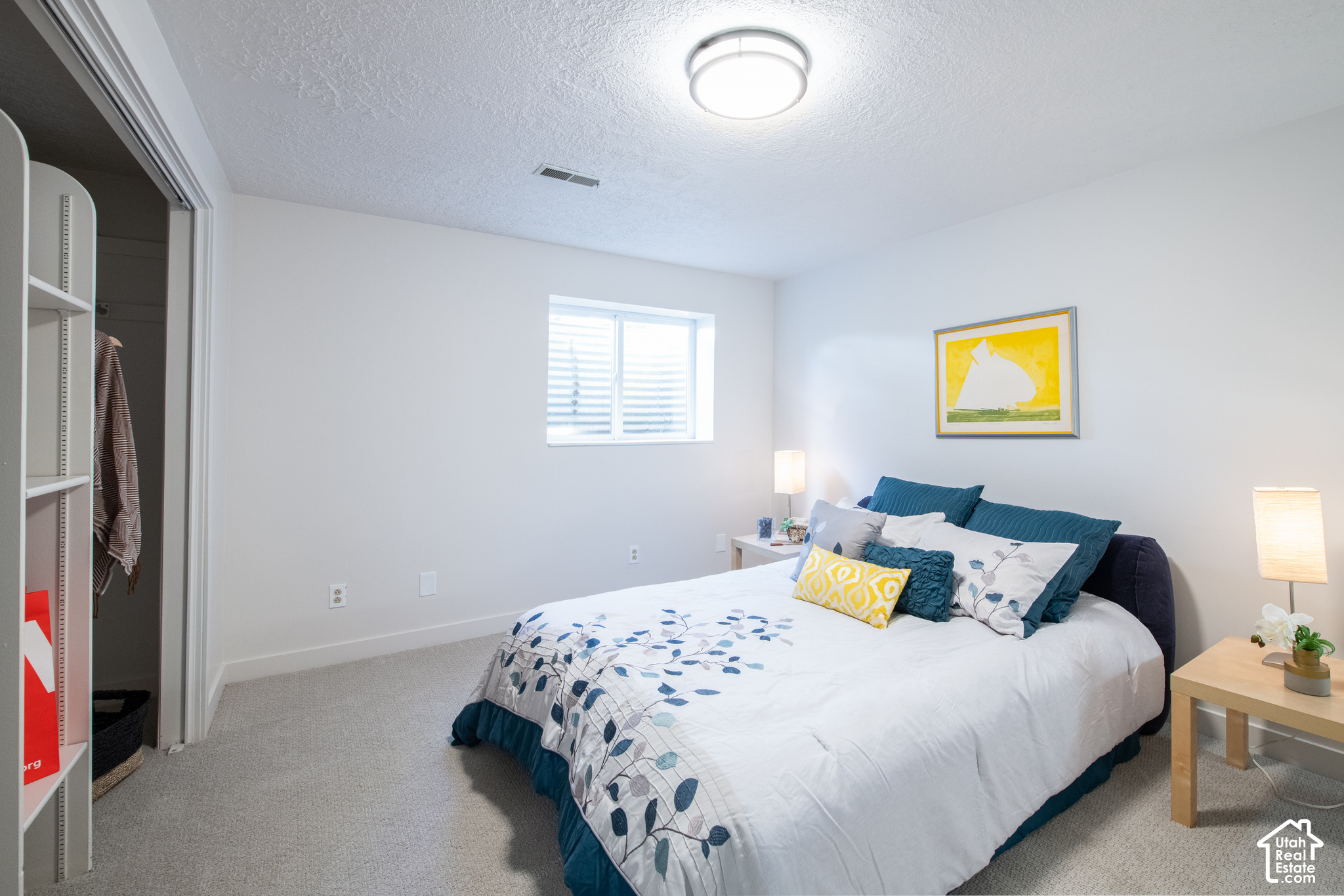 Furnished bedroom with white and blue bedding, teal pillows, and framed yellow artwork.