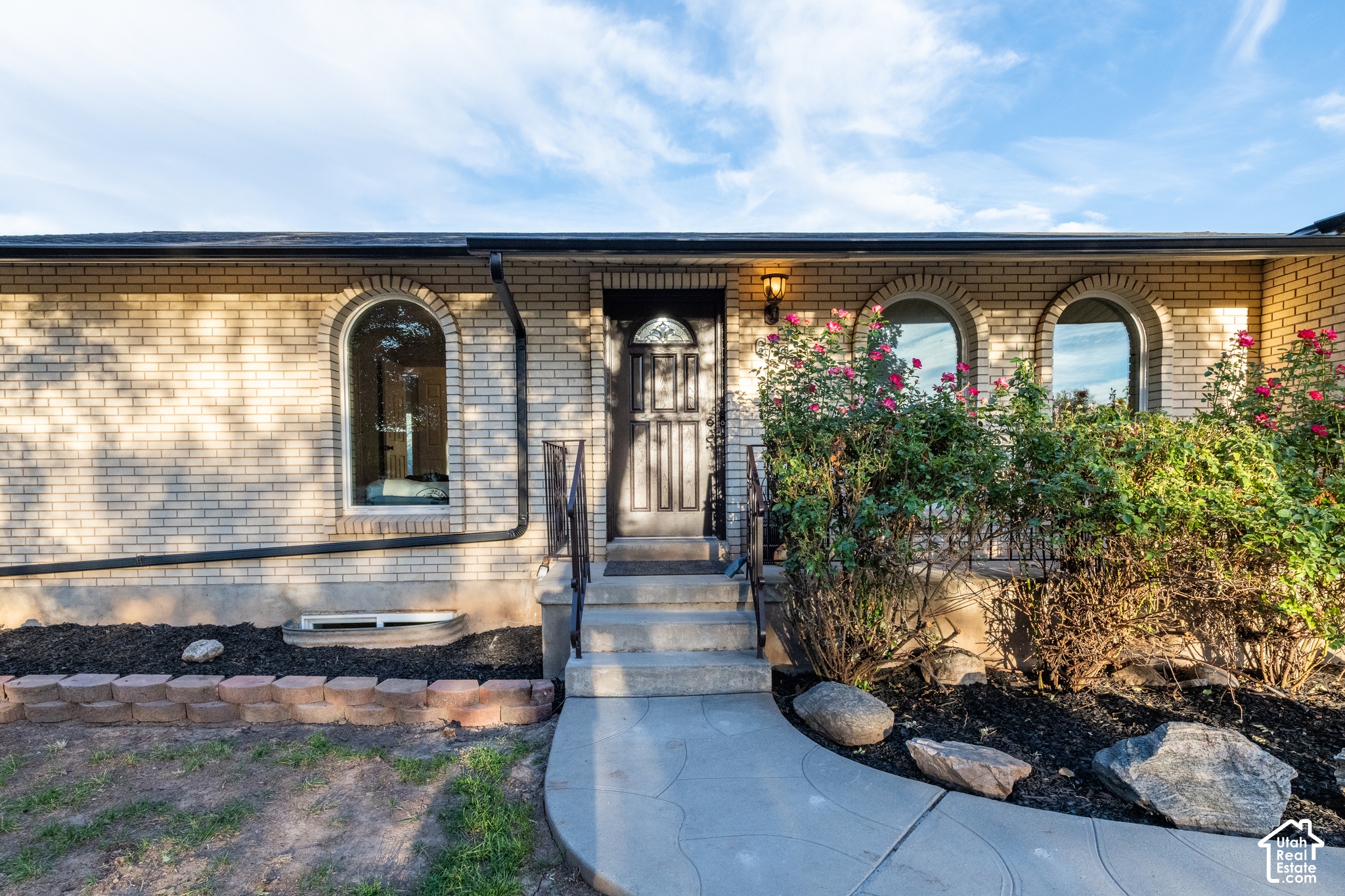 Light brick home entrance with arched windows, a curved walkway, and flowering rose bushes.