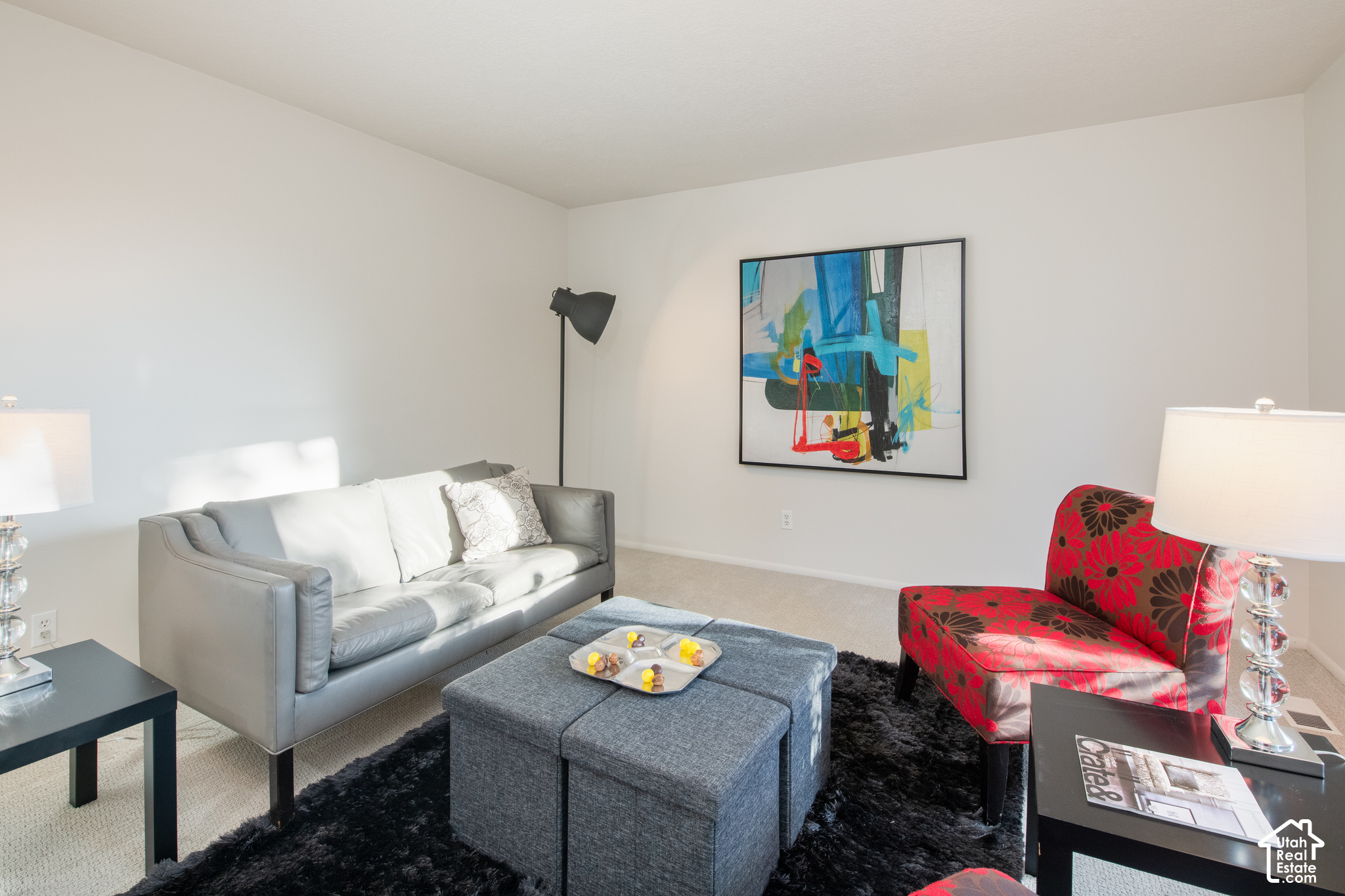 Staged living room with gray sofa, red floral armchair, blue ottoman, and abstract wall art.