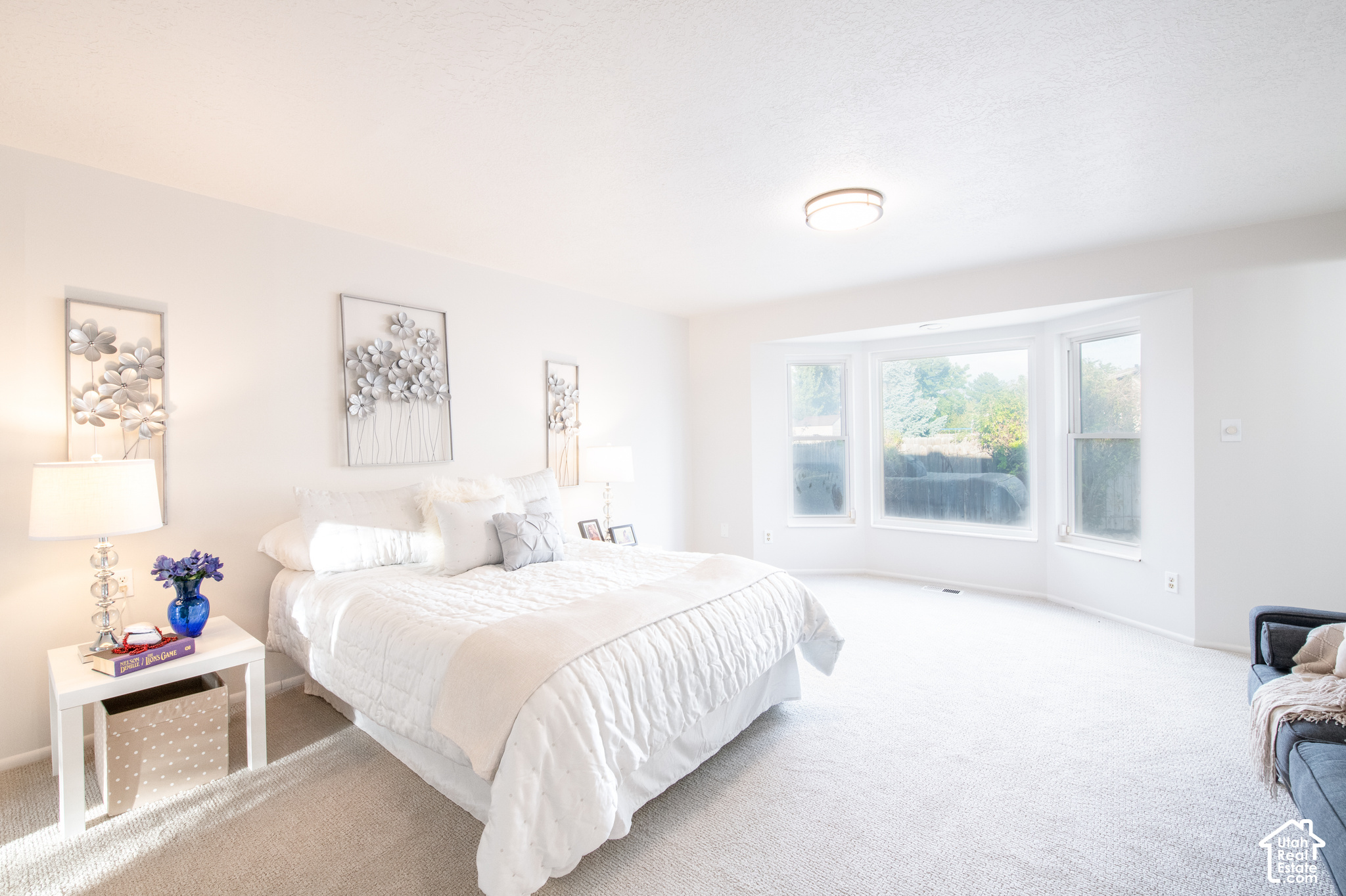 Bright bedroom with a white bed, floral wall art, and three large windows overlooking greenery.
