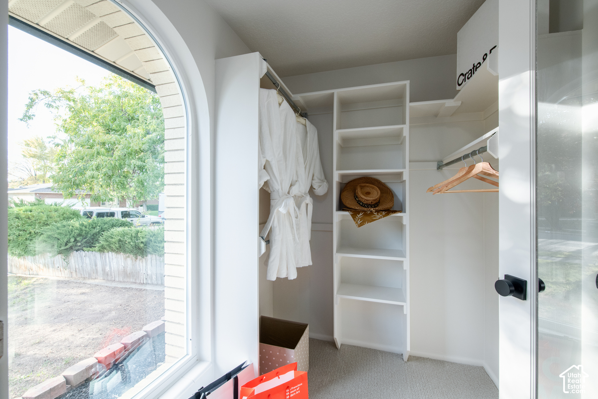 Walk-in closet with built-in shelving, hanging clothes, and an arched window overlooking a garden.
