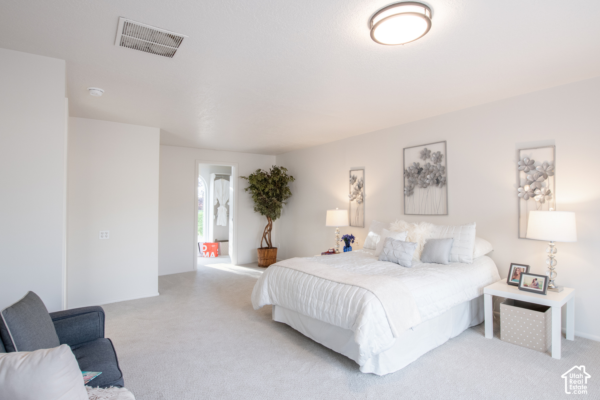 Staged bedroom with a white bed, floral wall art, and a potted tree.