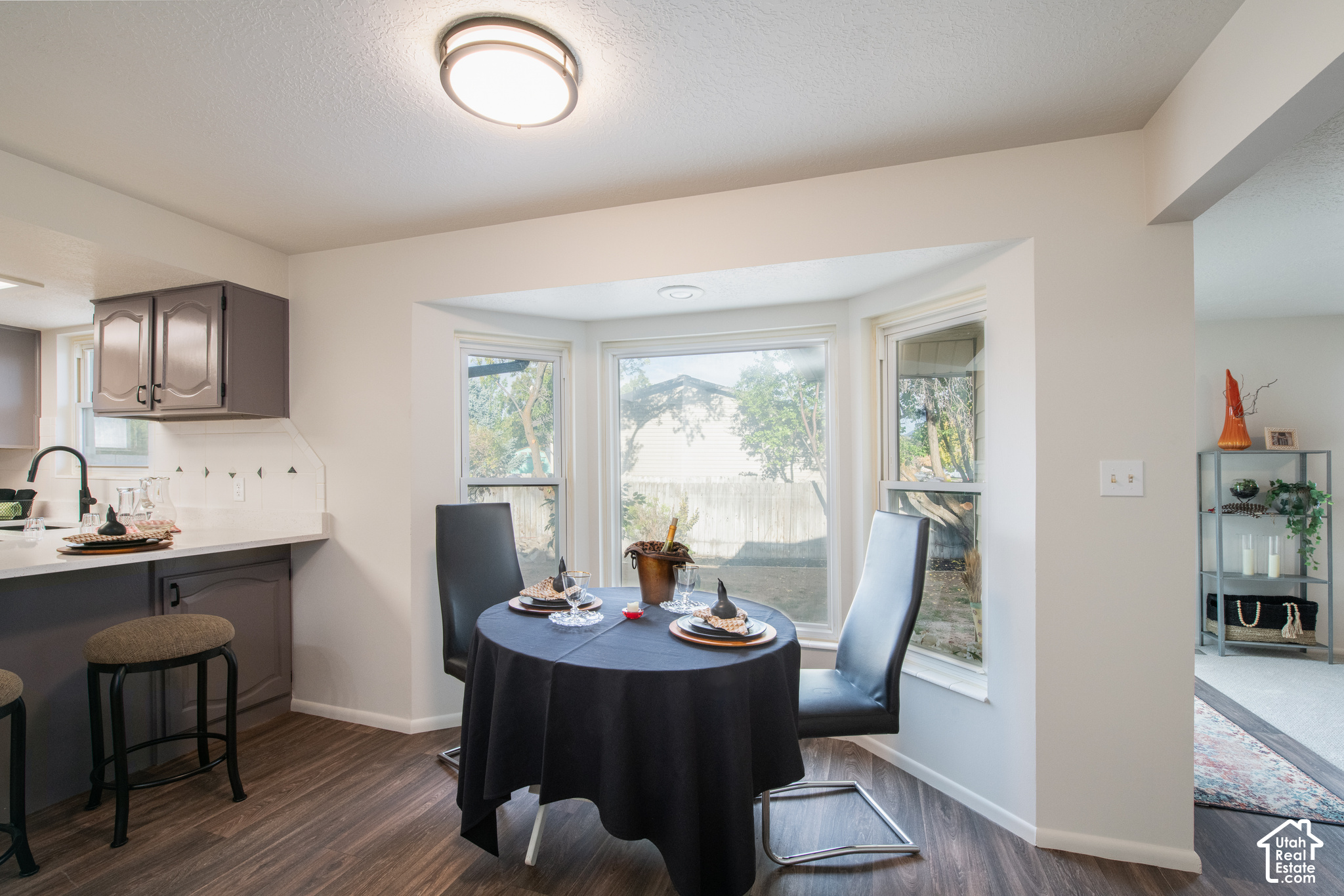 Dining nook with a table set for two beside large windows overlooking a fenced yard.