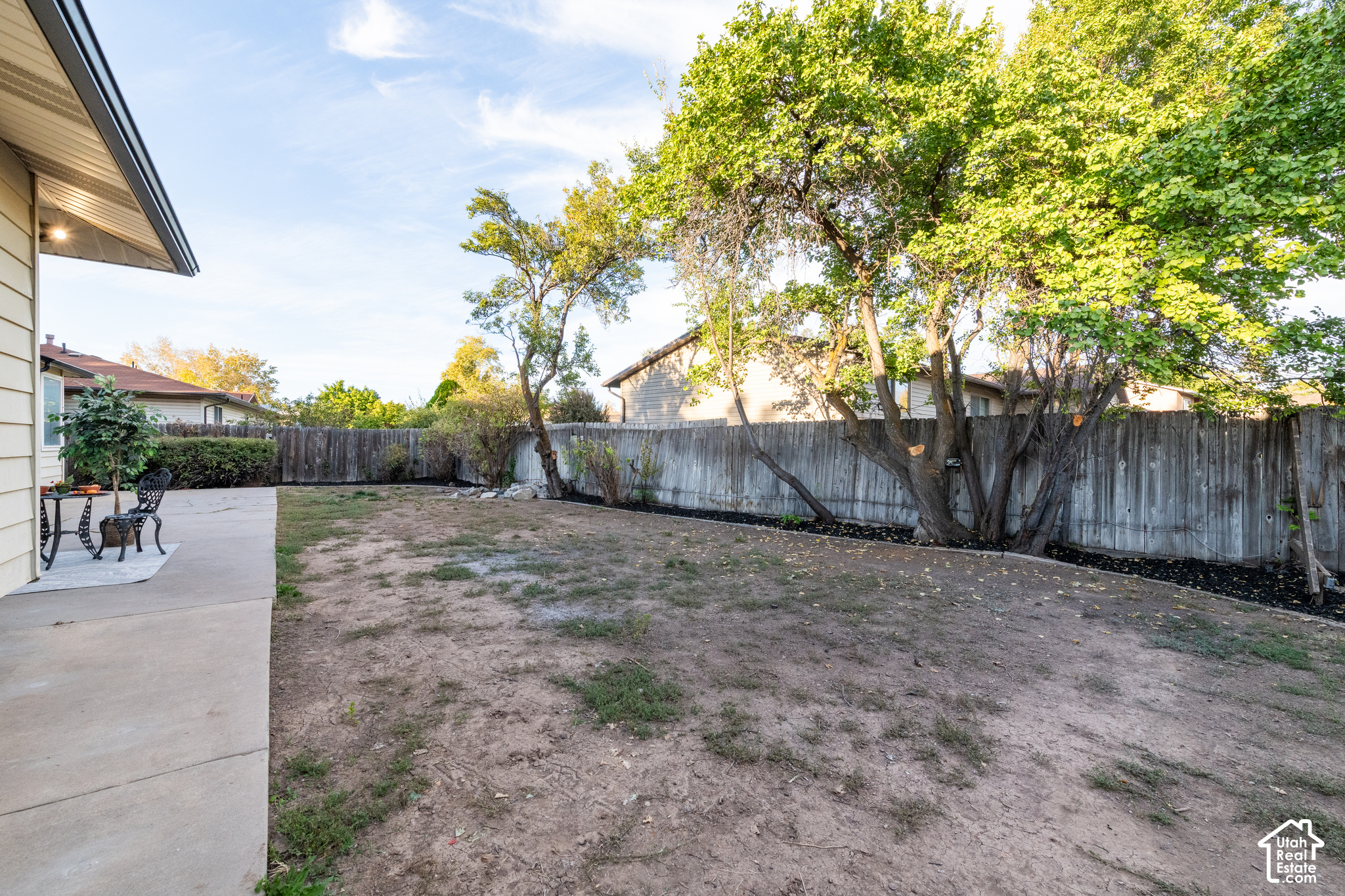 Fenced backyard with mature trees, a sparse lawn, and a concrete patio with outdoor furniture.