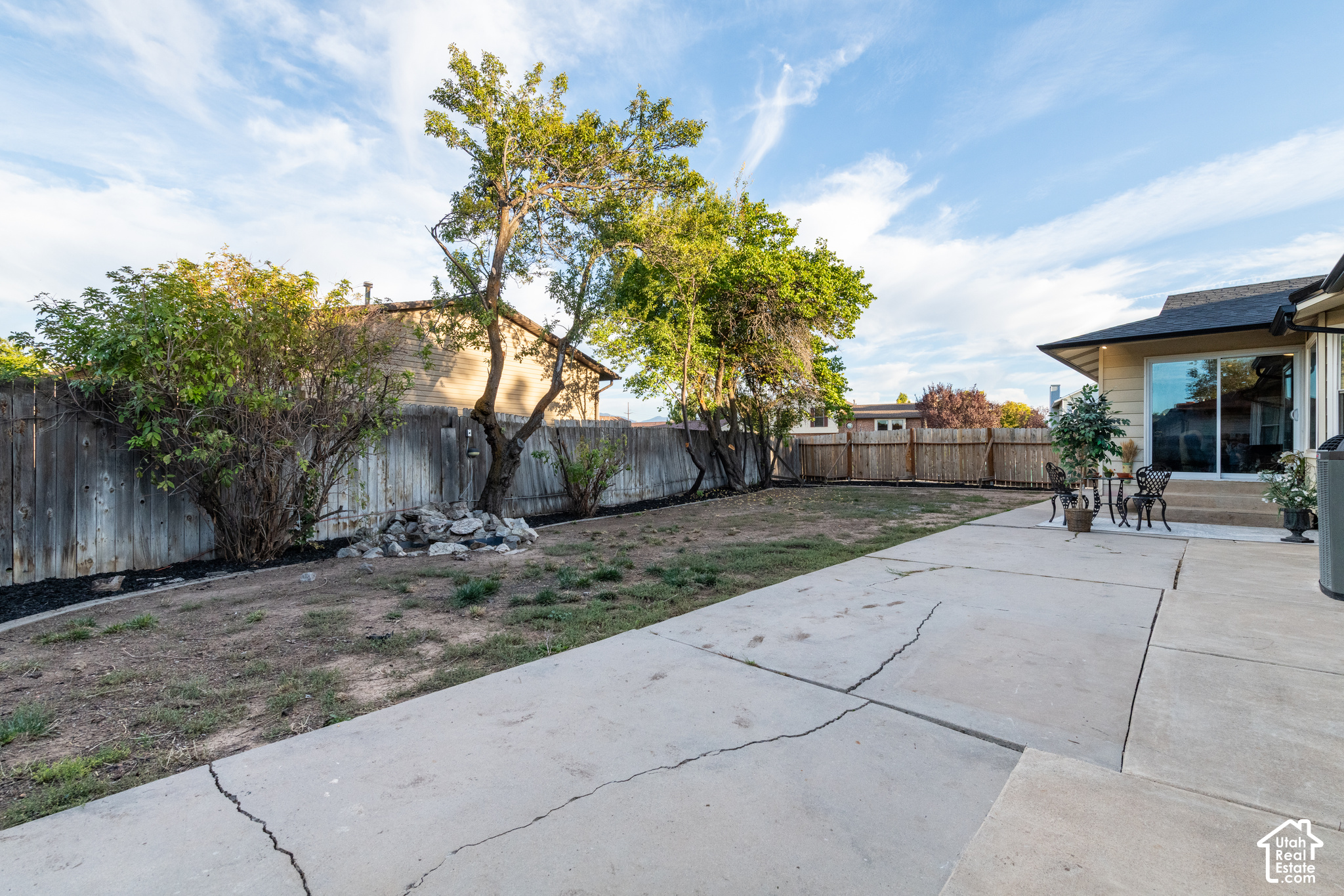 Fenced backyard with mature trees, patchy lawn, and a broad concrete patio beside the house.