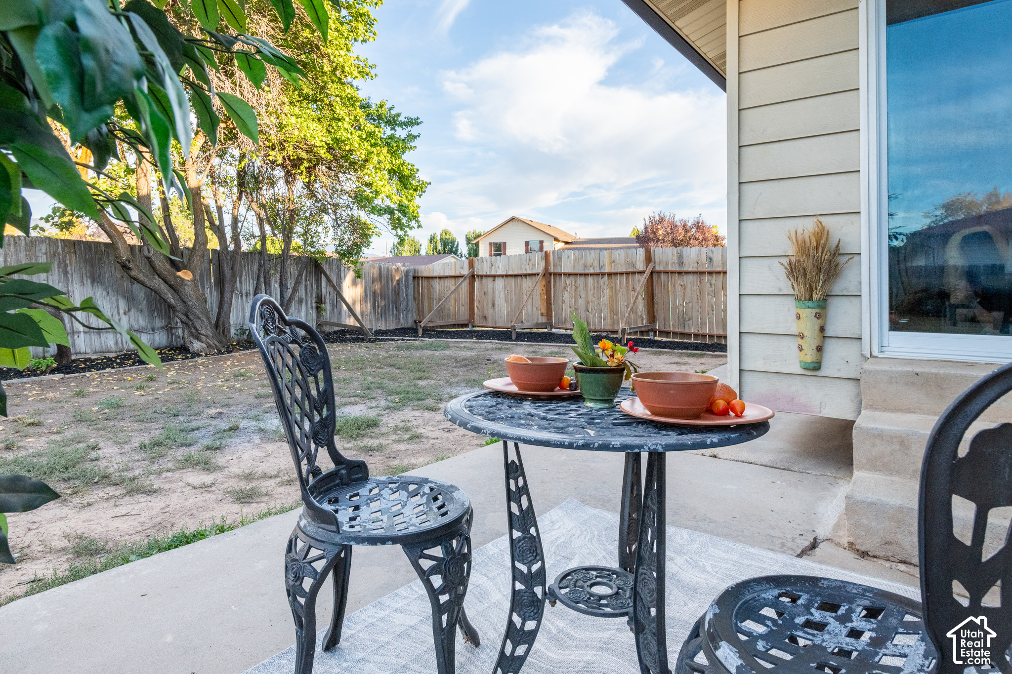 Furnished patio with a black metal bistro set beside a fenced backyard and mature shade tree.