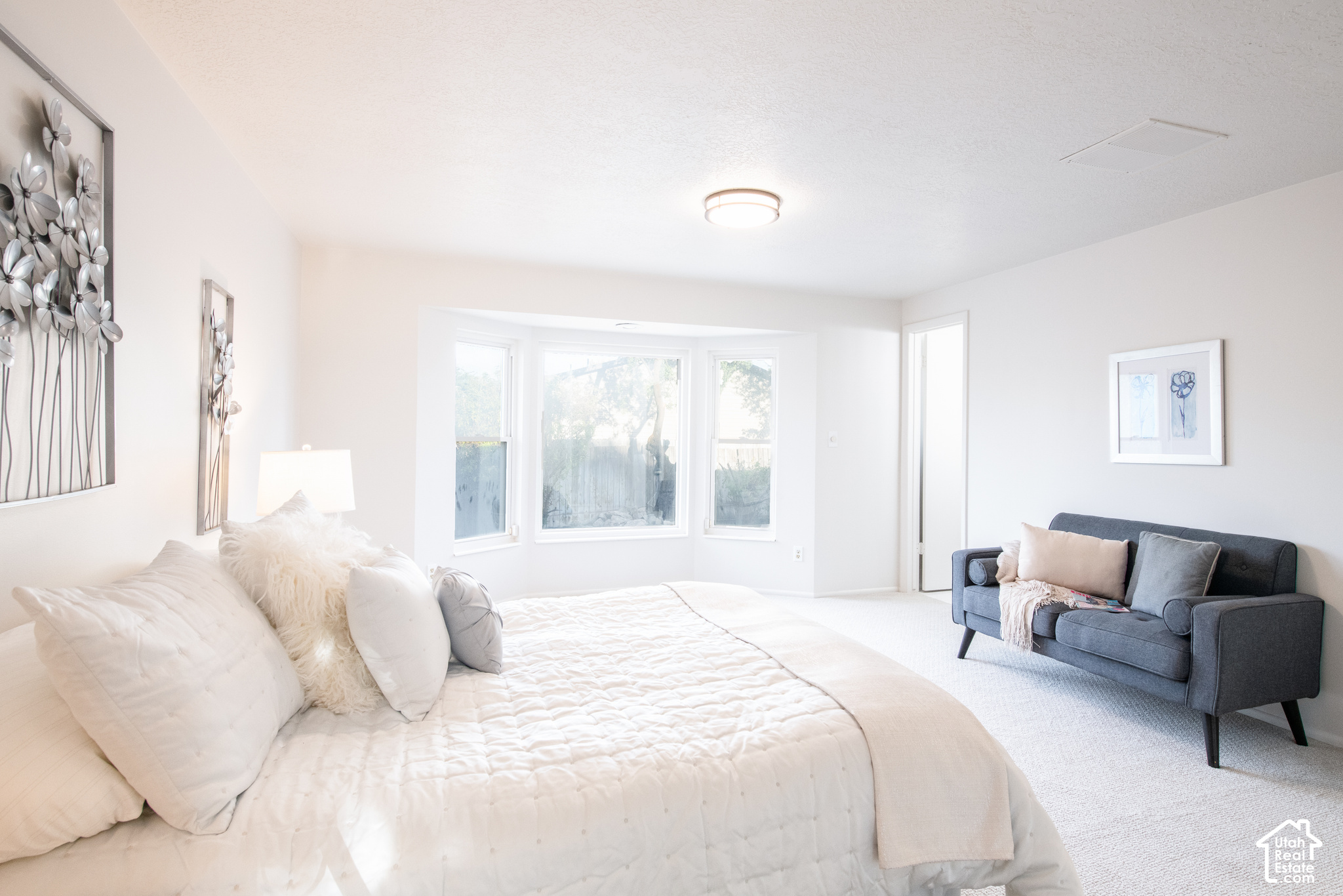 Bright bedroom with a large white bed, gray sofa, and three bay windows.