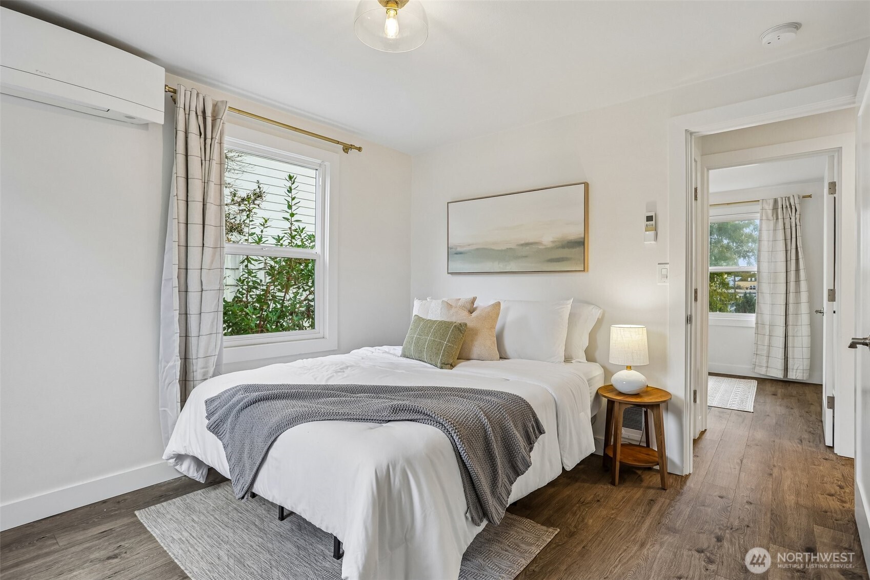 Staged bedroom with a white bed, gray throw, and window overlooking greenery.