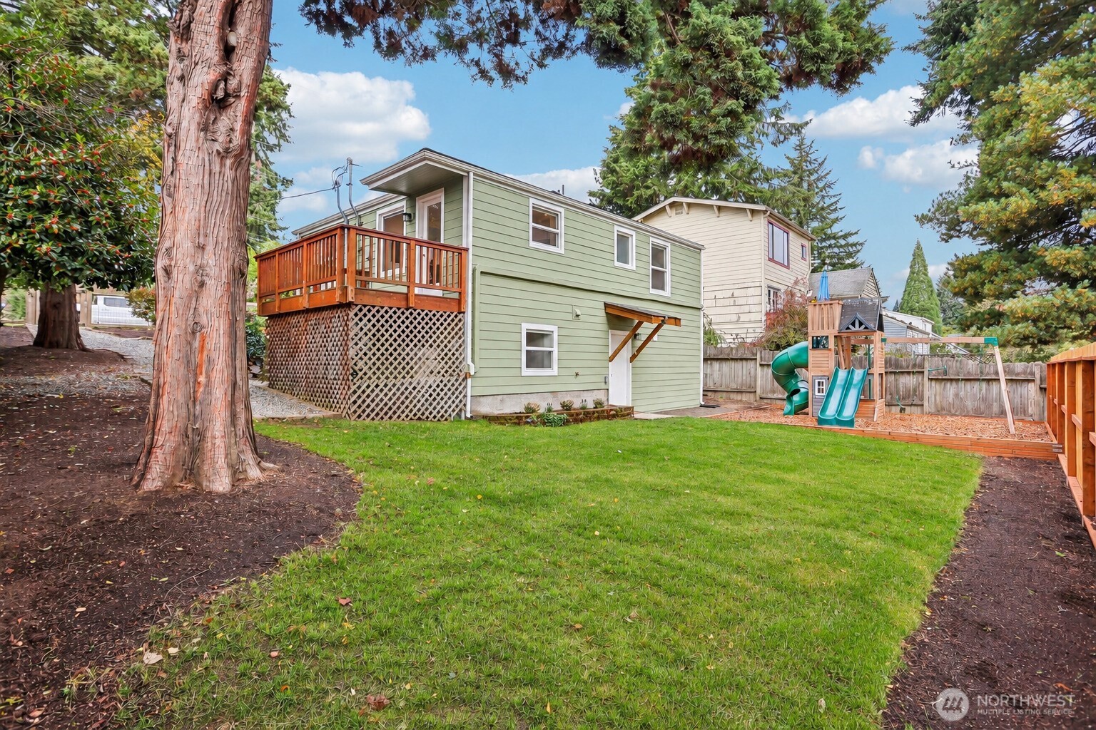 Green-sided home with a raised deck overlooking a fenced lawn and children's play structure.