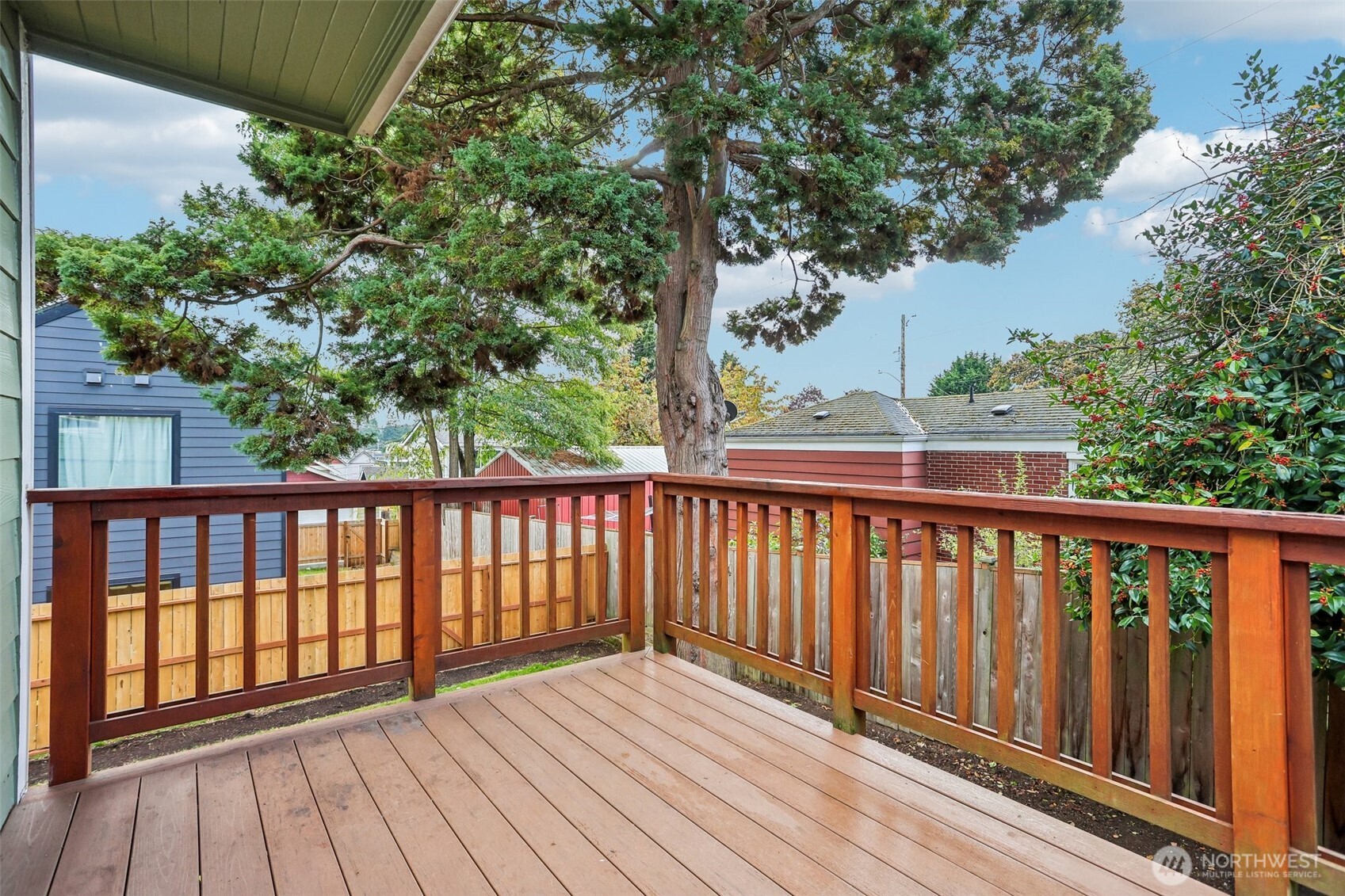 Wooden deck with reddish-brown railings overlooking a fenced yard and large evergreen tree.