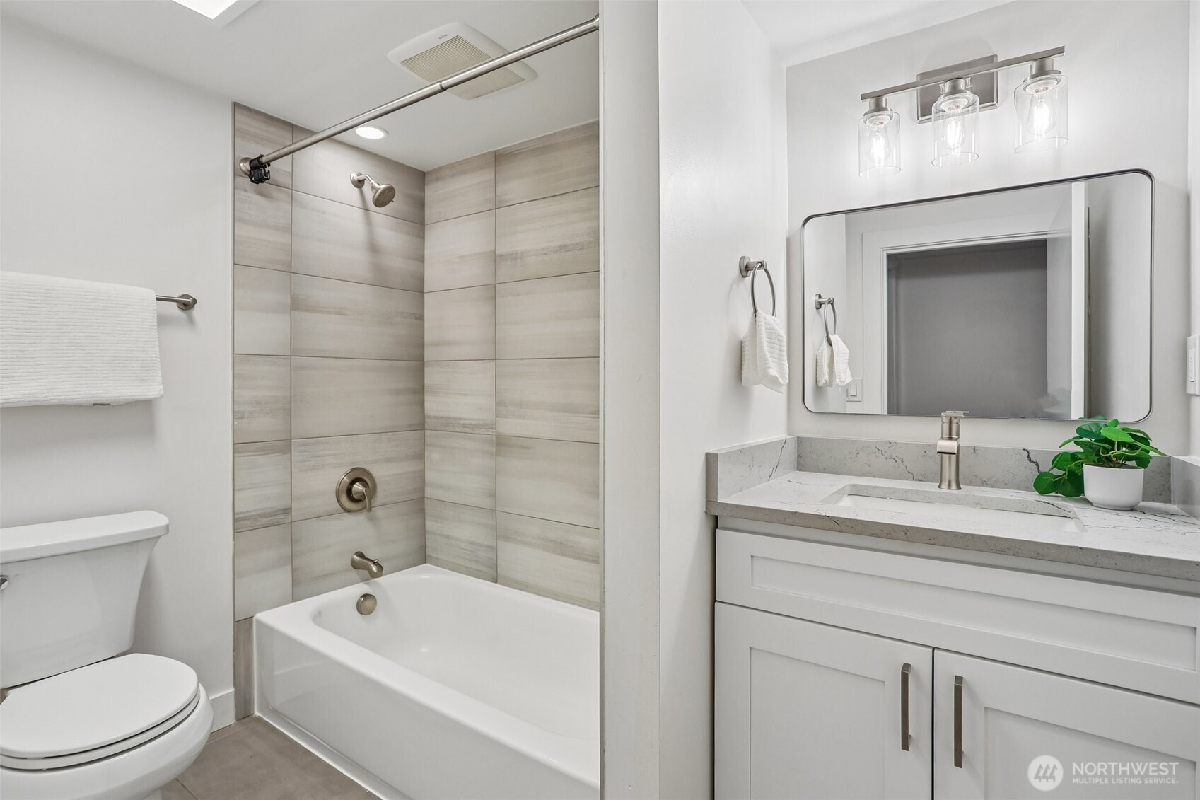 Bathroom with tiled tub and shower beside a white vanity with stone countertop and wide mirror.