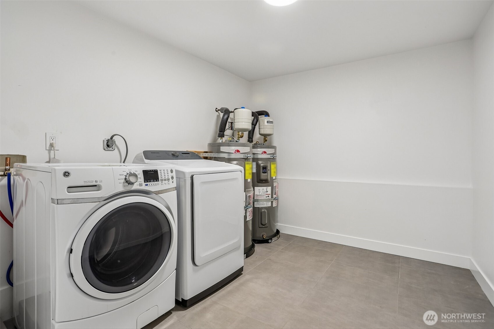 Laundry room with a front-loading washer and dryer, two water heaters, and gray tile flooring.