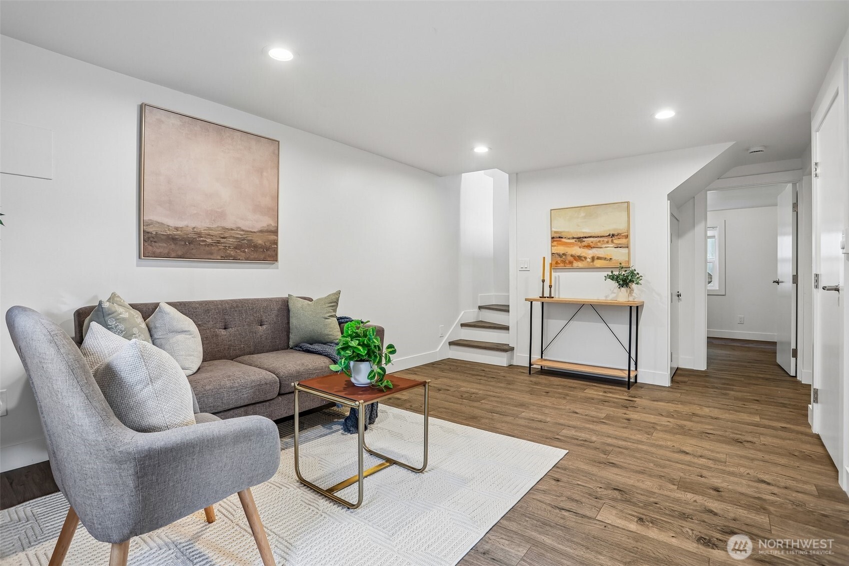 Staged living room with gray seating, warm wood flooring, and a short staircase.
