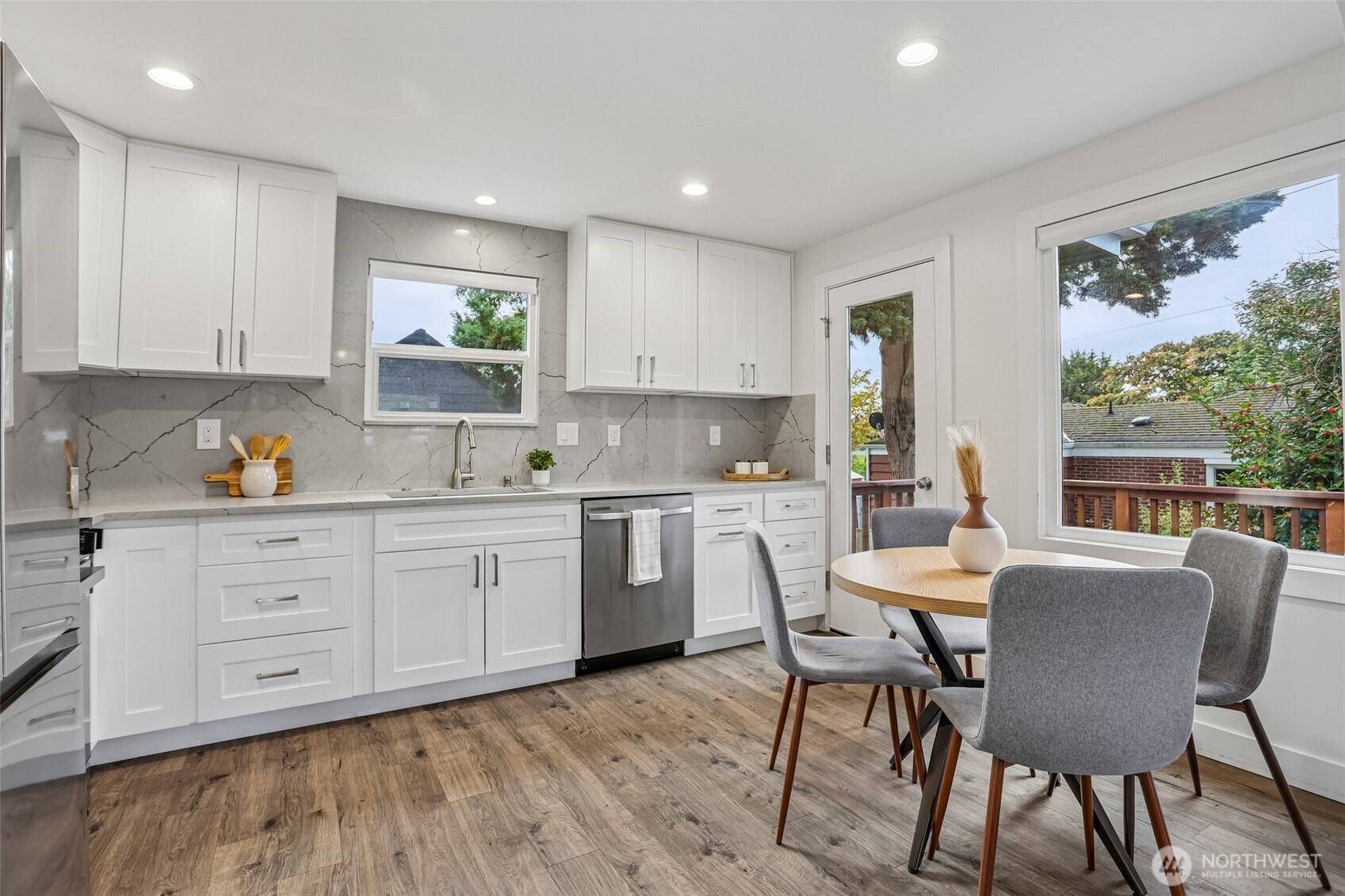 Bright kitchen with white cabinets, veined gray backsplash, and a dining table beside a large window.